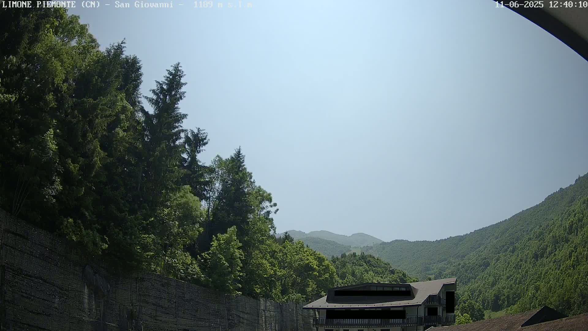 A partly sunny day reveals a mountainside covered in lush green trees, a building partially obscured by the view, and a high retaining wall in the foreground.
