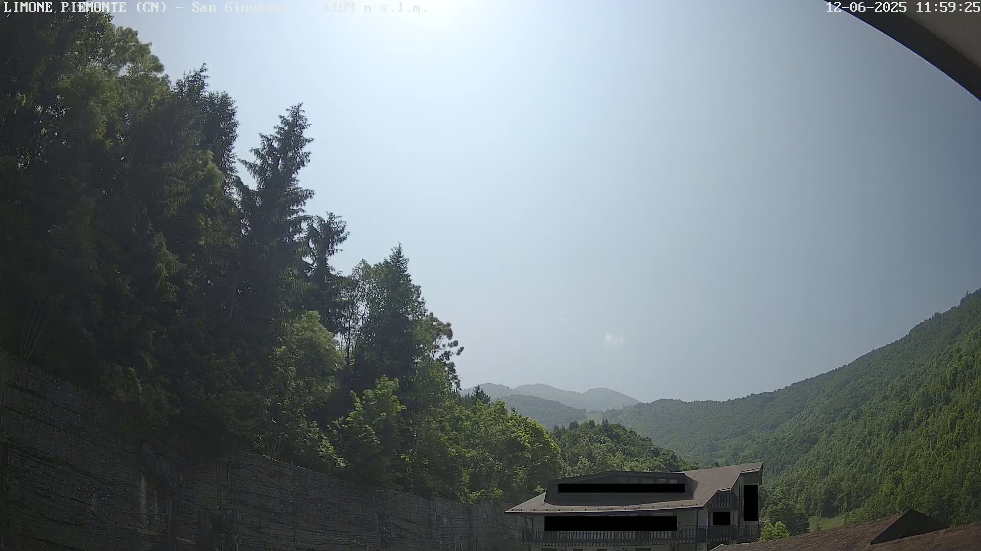 A hazy, sunny day reveals a mountainside covered in lush green trees, with a building partially visible near the bottom.