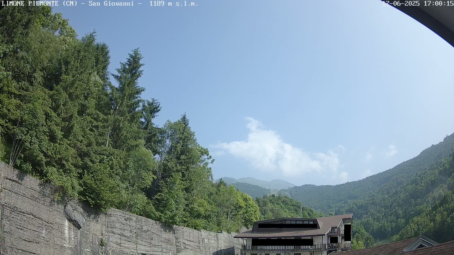 A mostly clear blue sky with a few puffy clouds overlooks a lush green mountainside and forested area, partially obscured by a retaining wall and a building.