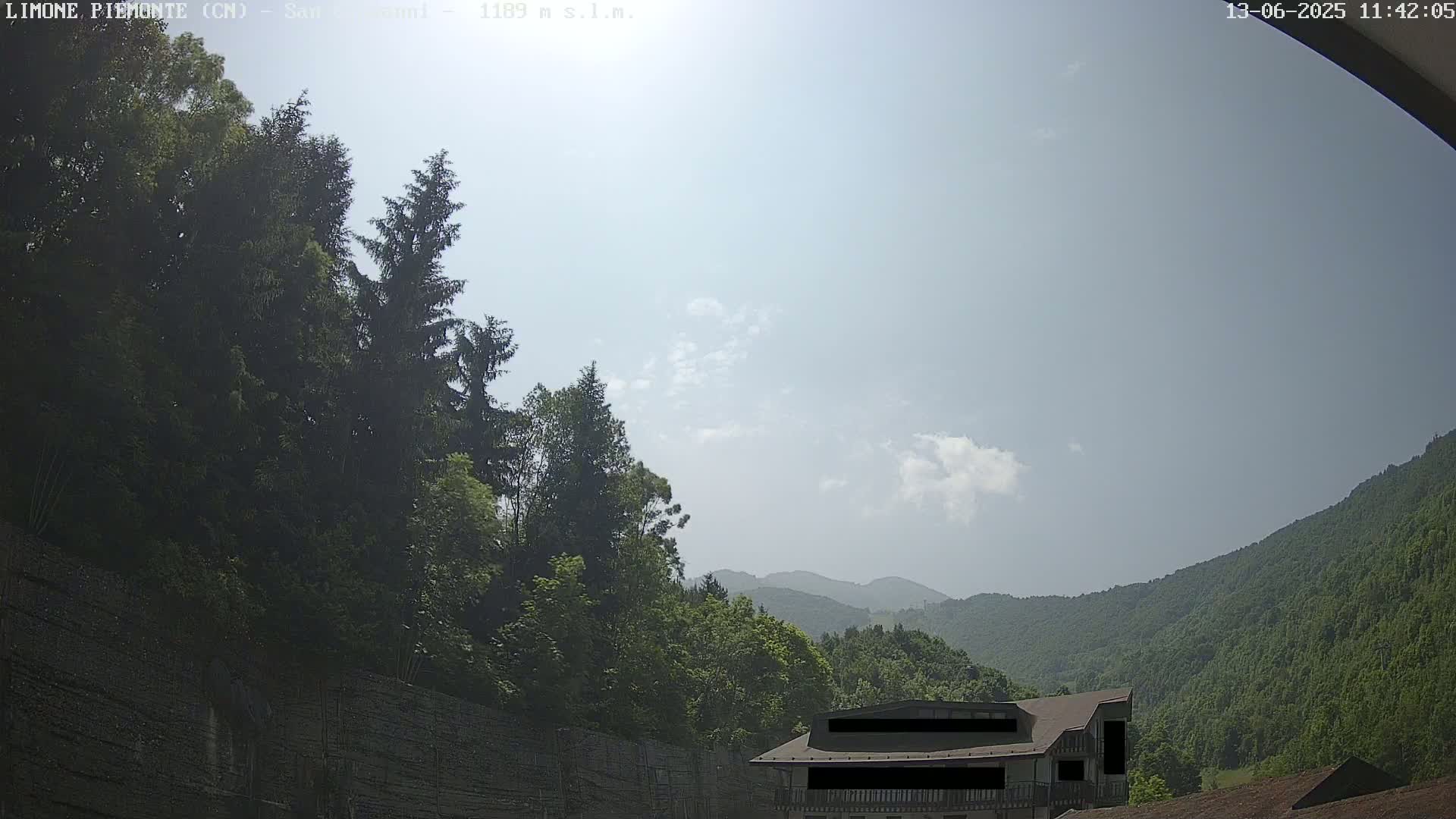 A partly cloudy, sunny day reveals a mountain valley with lush green trees and a building partially obscured by a wall.