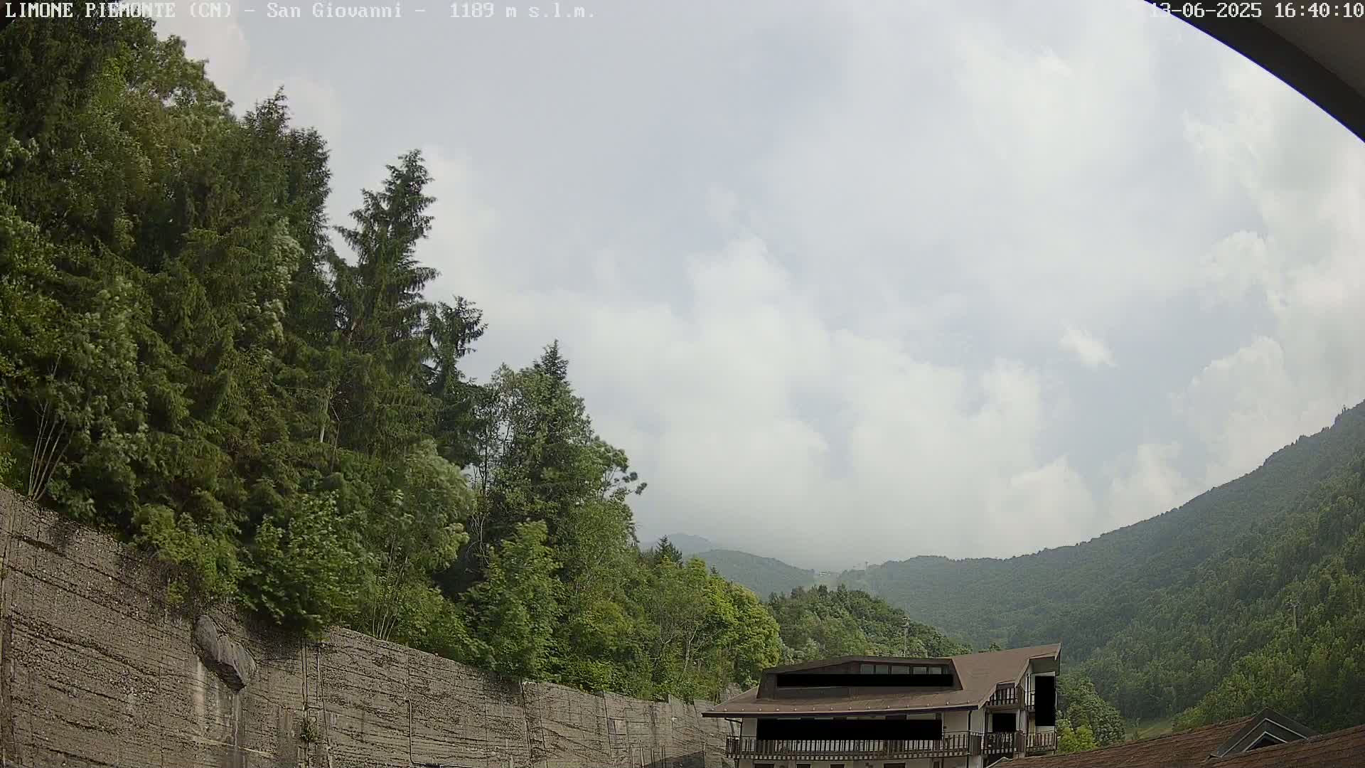 A partly cloudy sky overlooks a mountainous landscape with lush green trees, a building nestled among the greenery, and a retaining wall at the foreground.