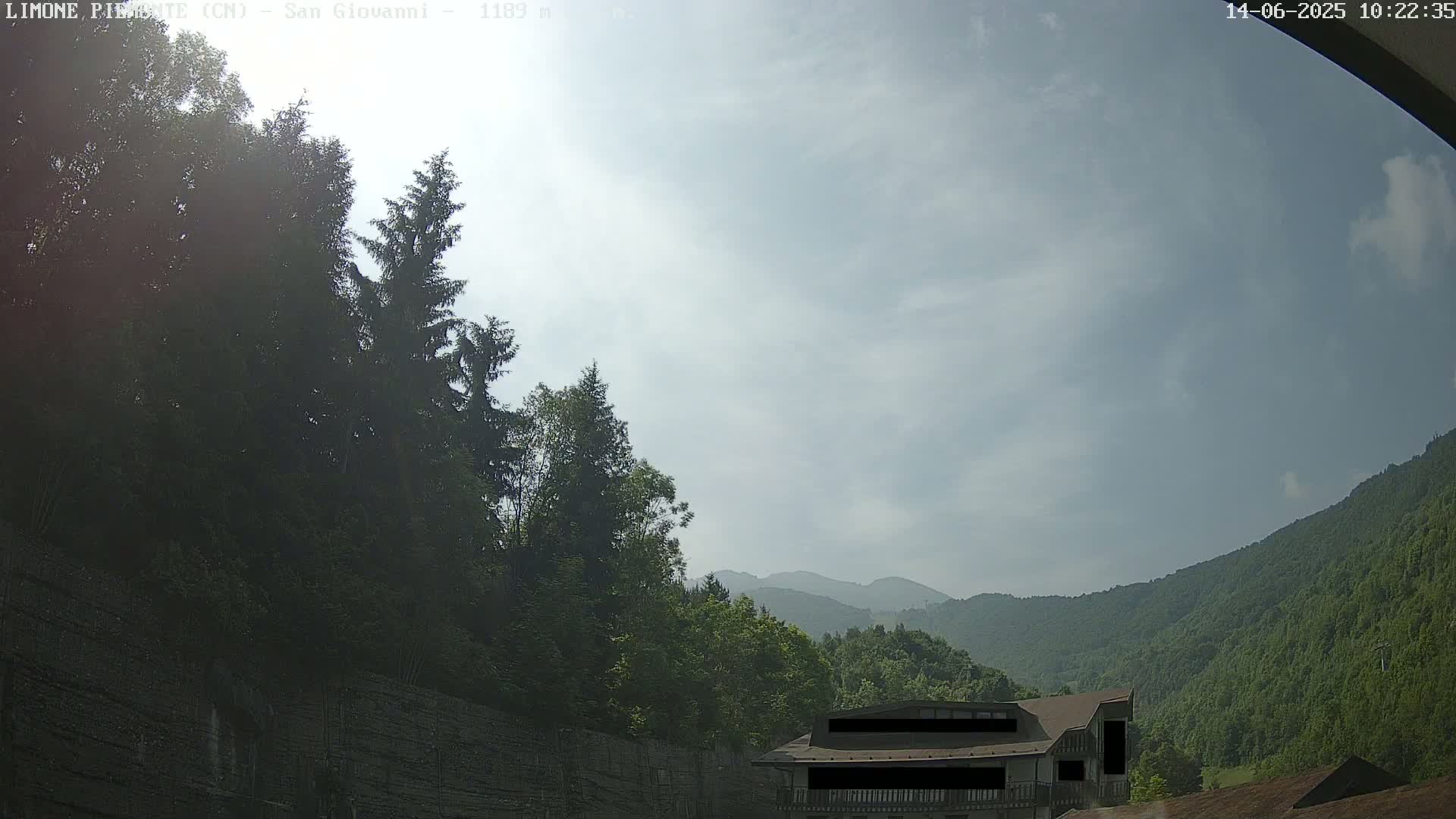 A partly cloudy sky overlooks a mountainside with green trees and a building partially obscured by a wall.