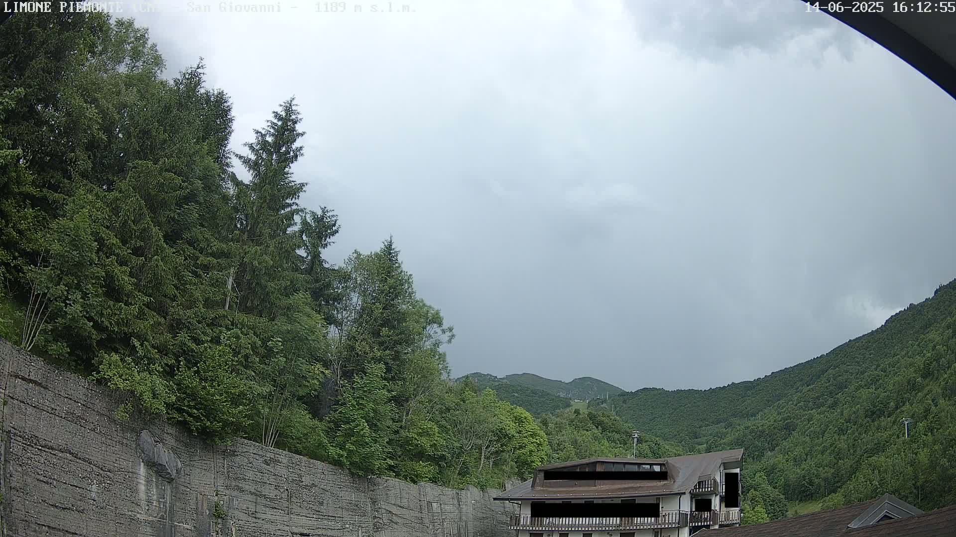 A cloudy sky hangs over a mountainous landscape of lush green trees and a building partially obscured by foliage, with a retaining wall in the foreground.