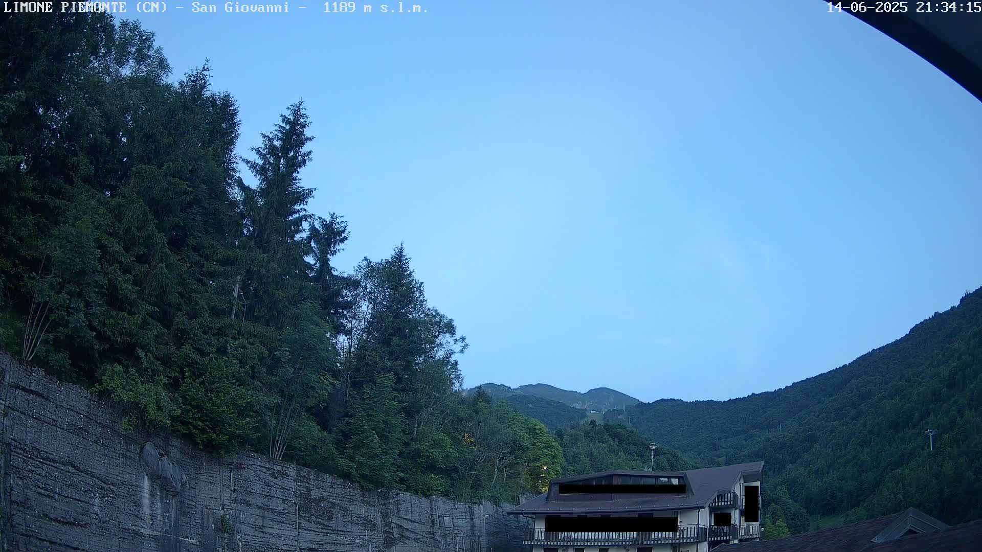 A cloudy sky hangs over a mountainous landscape of lush green trees and a building partially obscured by foliage, with a retaining wall in the foreground.