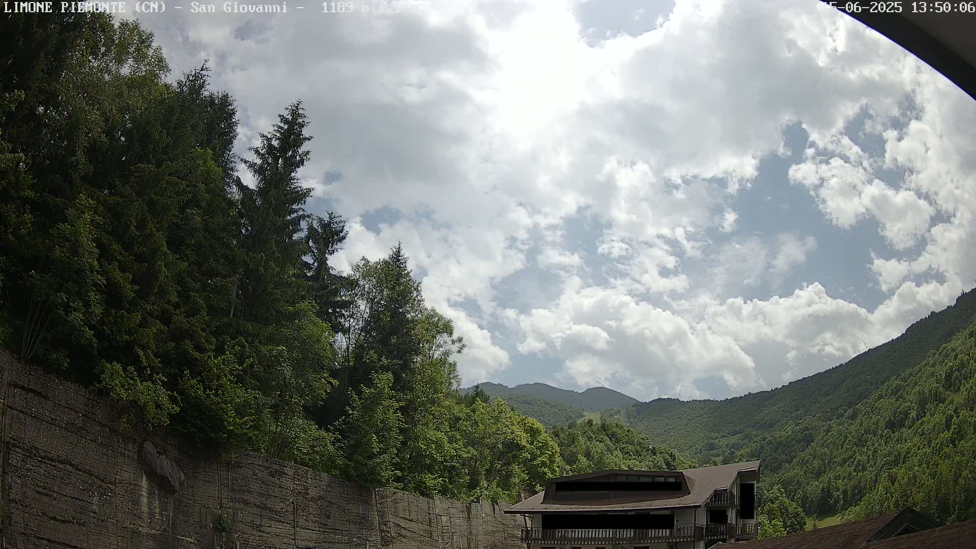 A partly cloudy sky overlooks a mountainous landscape, with lush green trees and a building partially visible near a stone wall.