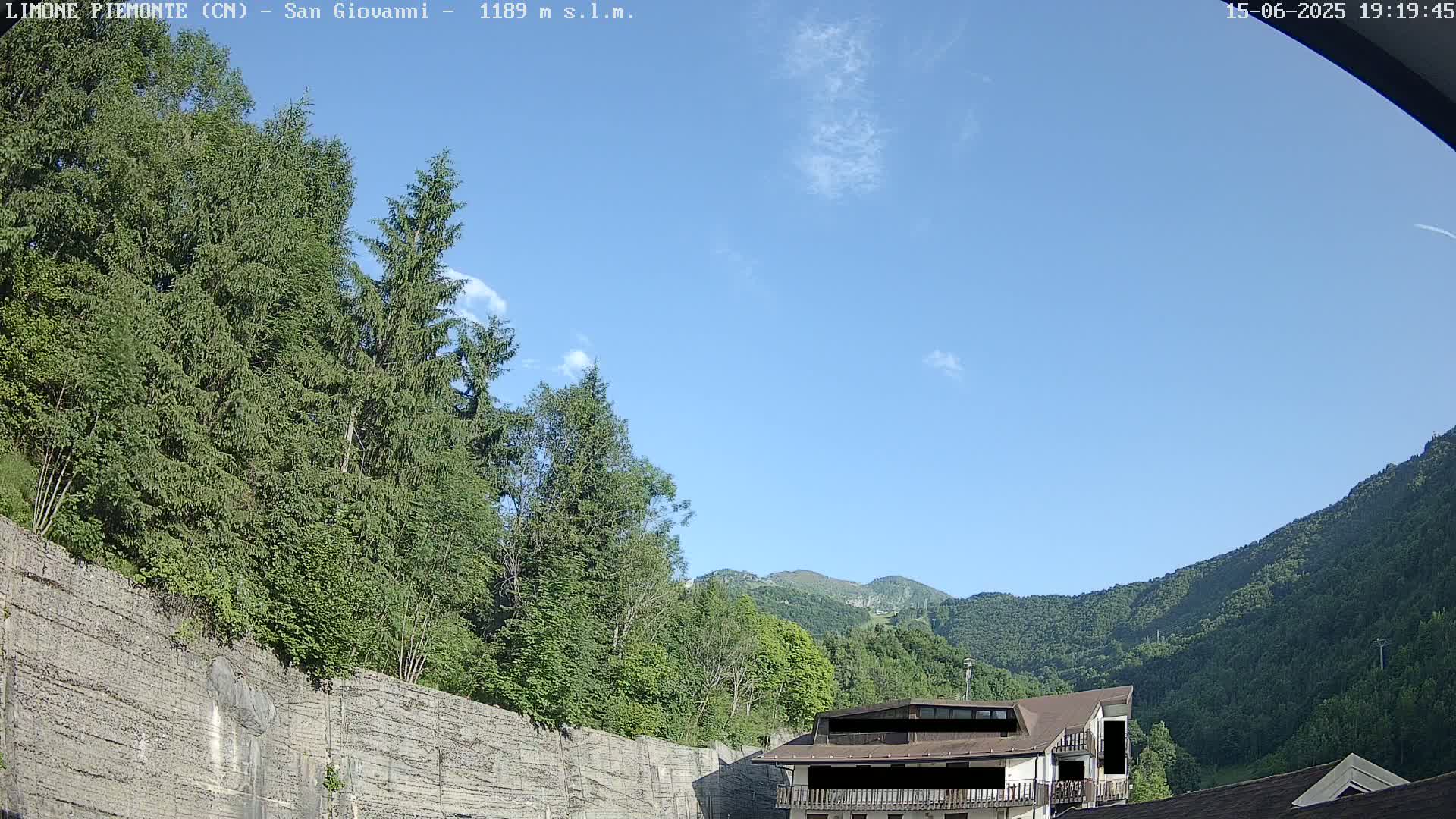 A mostly clear blue sky overlooks a mountainside with lush green trees, a stone retaining wall, and a building with a brown roof.