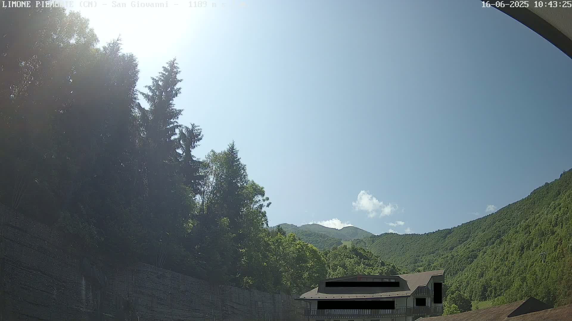 A sunny day reveals a partially visible building nestled among lush green trees and mountains under a mostly clear sky.