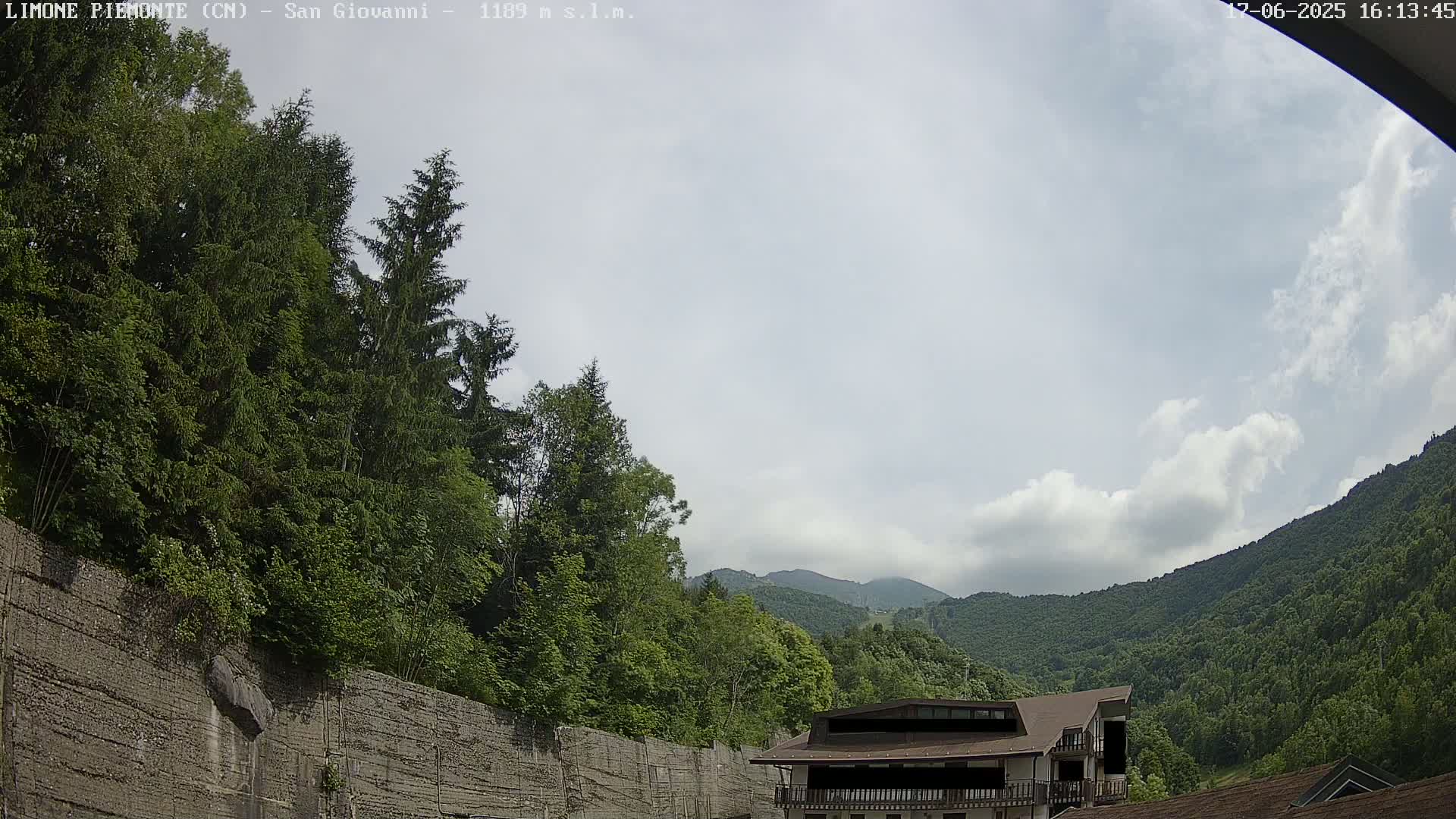A partly cloudy sky overlooks a mountainous landscape of lush green trees, a building, and a tall retaining wall.