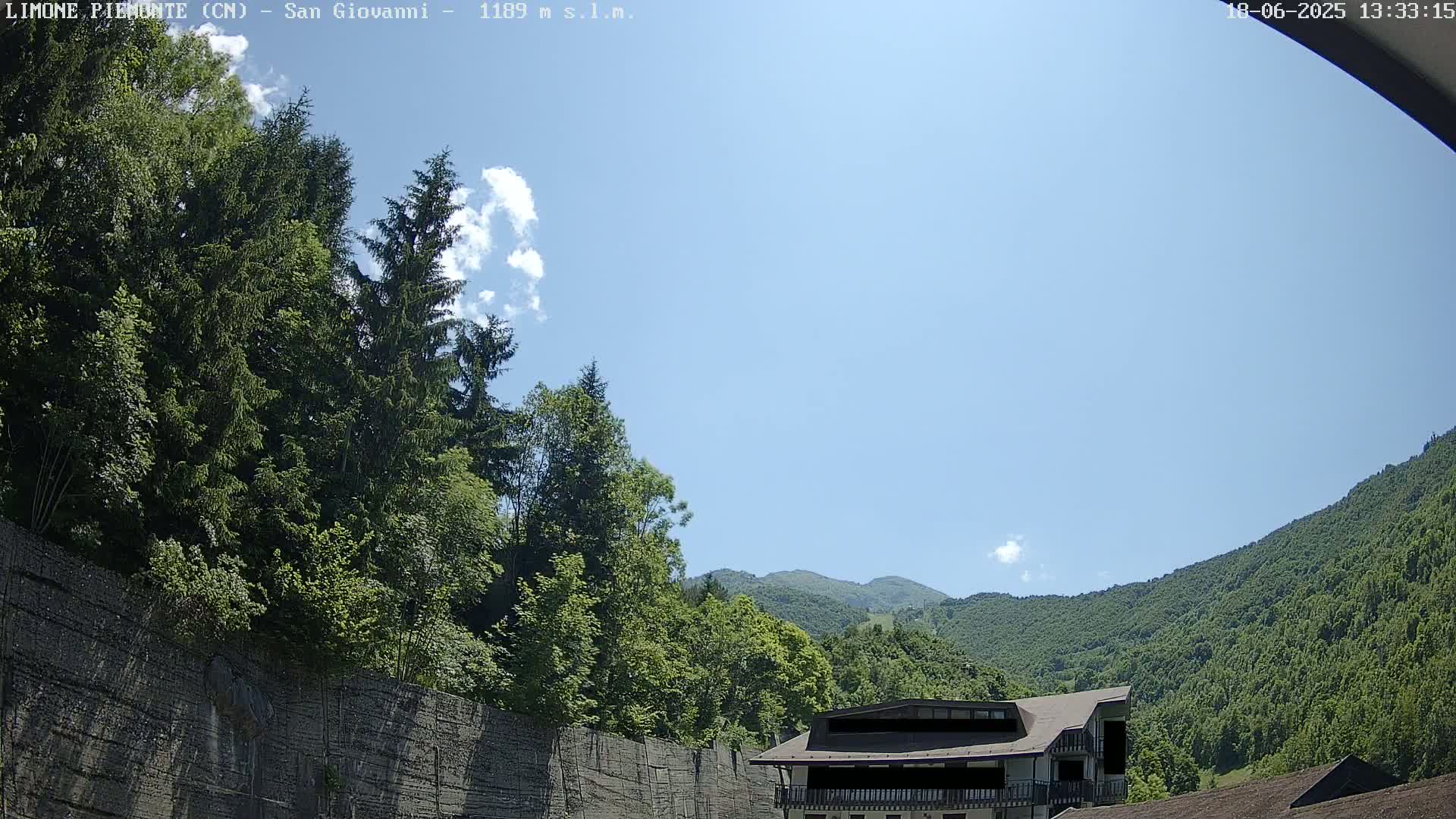 A mostly sunny sky is above a building nestled in a lush green mountainside, partially obscured by a tall retaining wall.