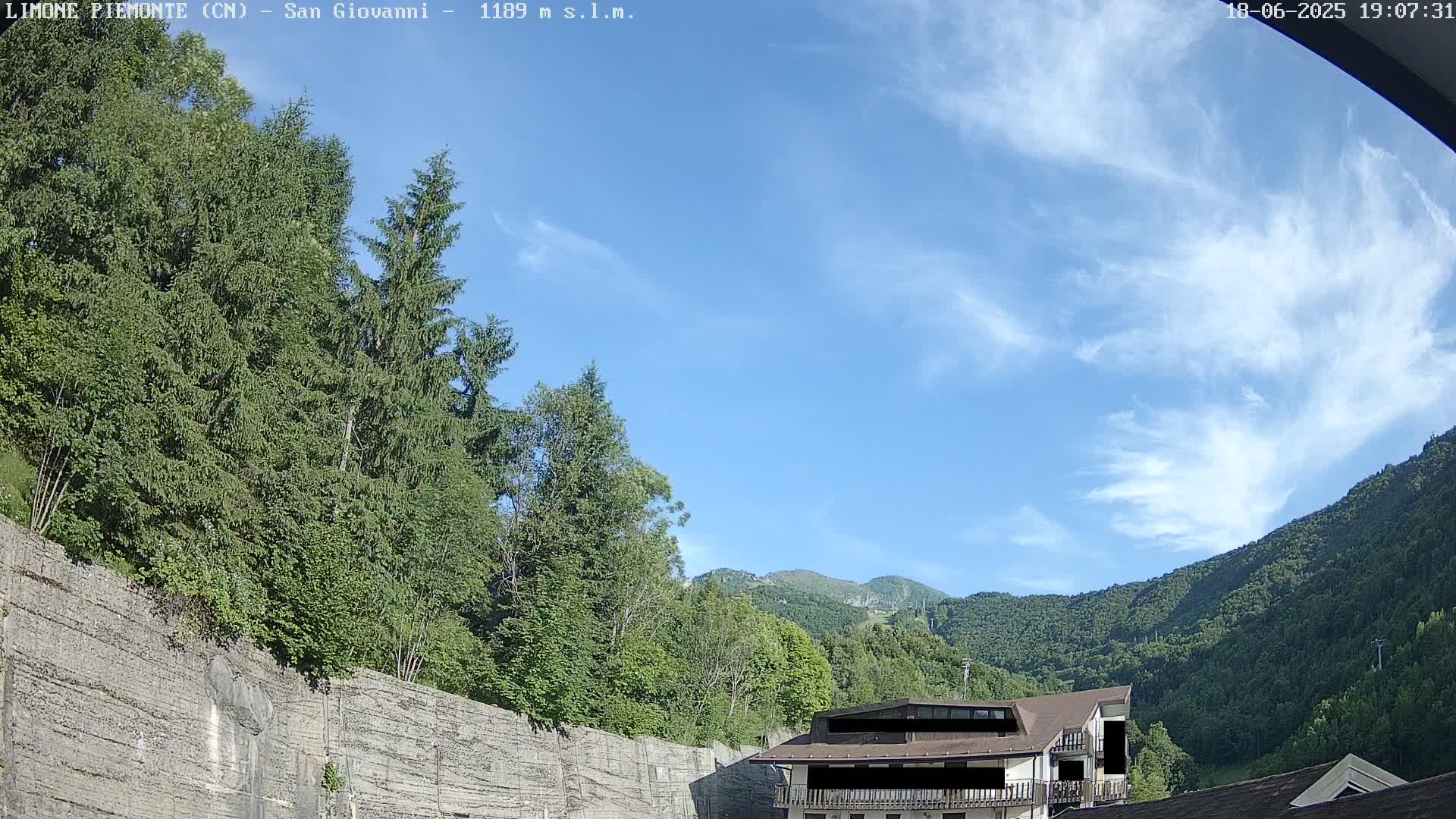 A partly cloudy blue sky overlooks a mountainous landscape of green trees, a building, and a large stone retaining wall.