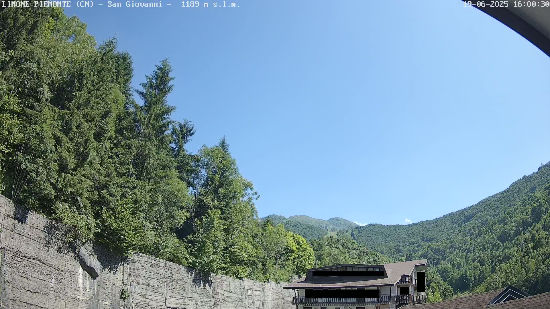 A sunny day reveals a view of lush green trees and mountains, with a building partially visible nestled against a concrete retaining wall.