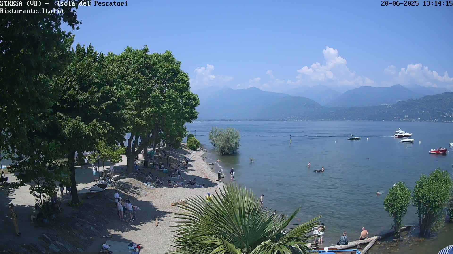 A sunny day on a beach with people sunbathing and swimming in calm water,  boats on the lake, and mountains in the background.