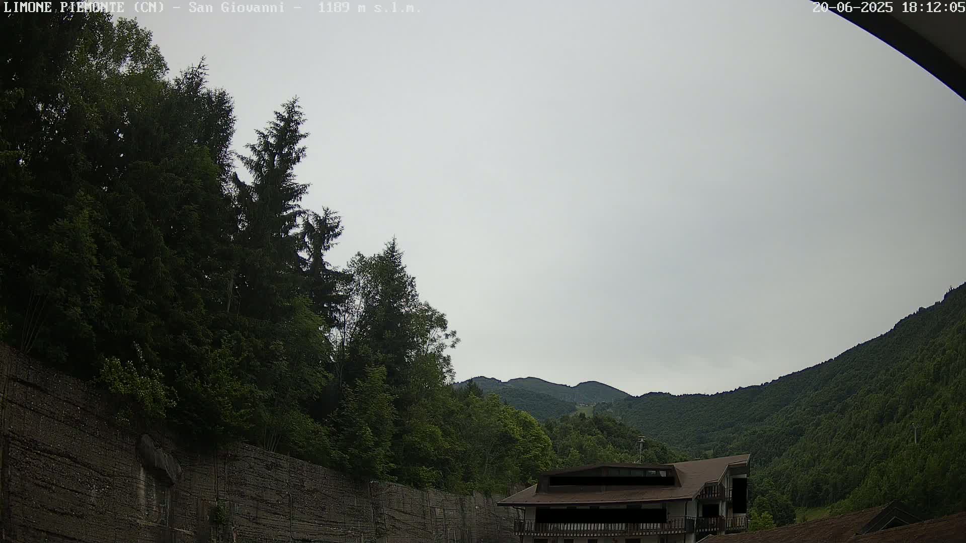 A cloudy sky overlooks a mountainside covered in lush green trees, partially obscured by a tall, dark retaining wall and a building with a brown roof.