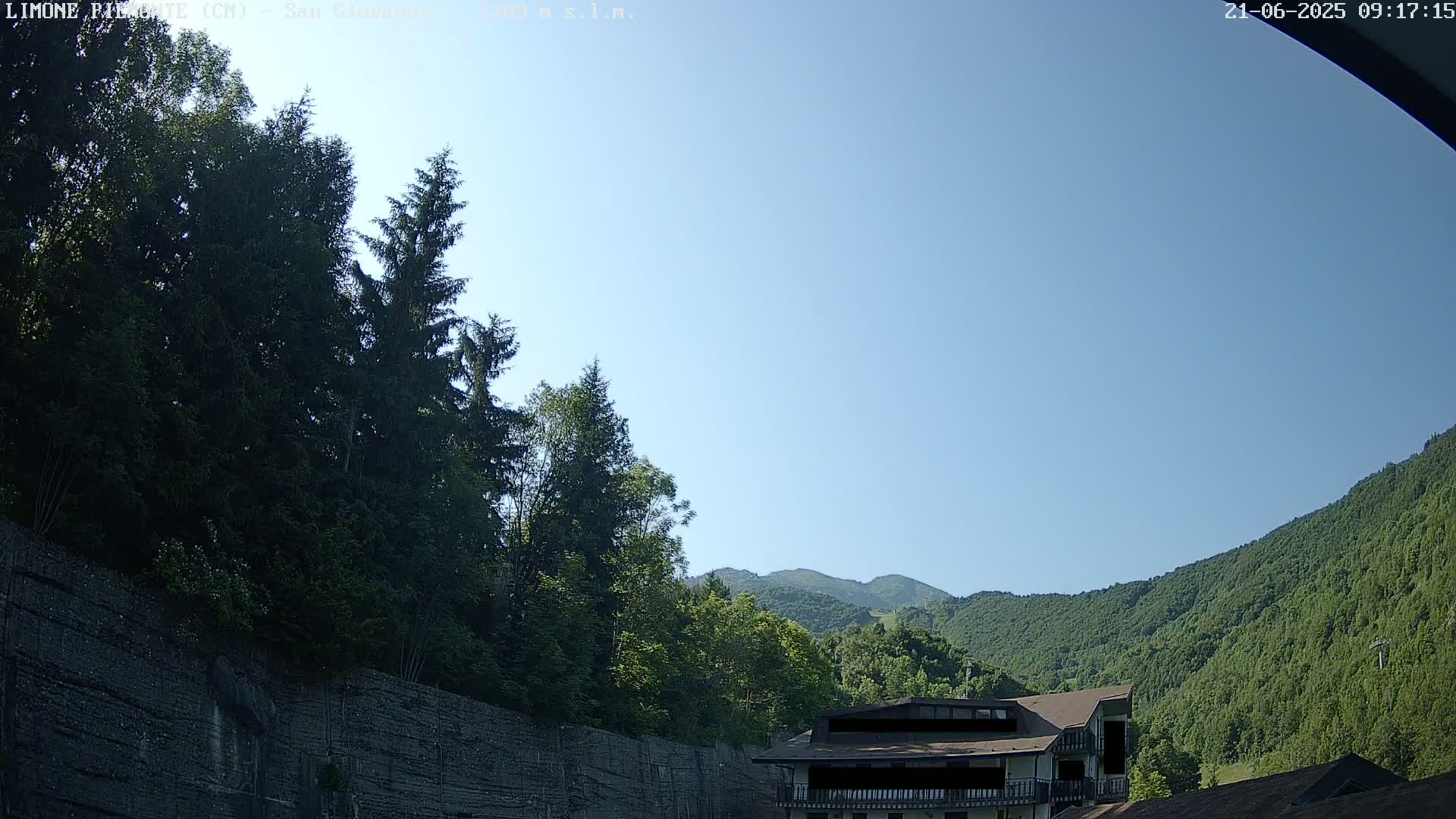 A clear blue sky is visible above a mountainside covered in lush green trees and a building partially obscured by foliage.