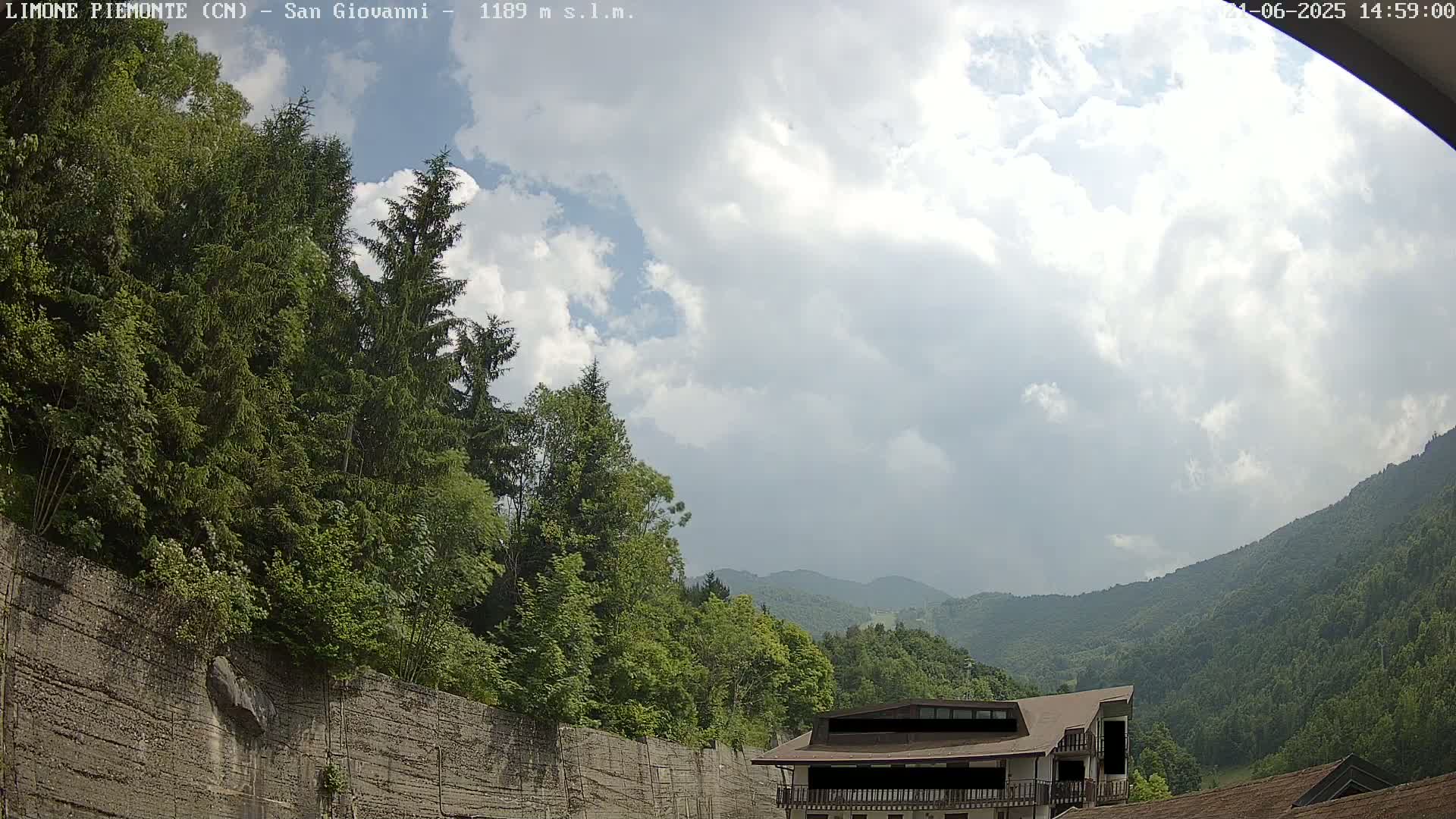 A partly cloudy sky overlooks a mountainous landscape of green trees and a building, with a retaining wall in the foreground.