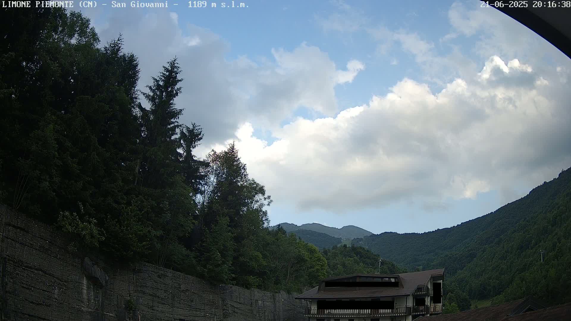 A partly cloudy sky overlooks a mountainside landscape with green trees, a building, and a tall stone wall.