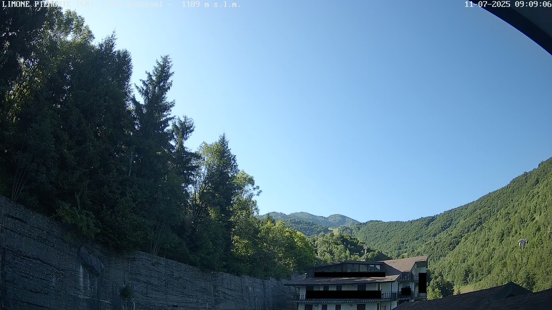 A clear blue sky is visible above a green mountainside partially obscured by trees and a building.