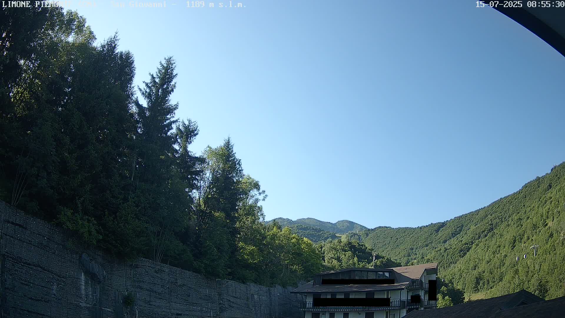 A clear blue sky overlooks a mountainside covered in lush green trees, with a building partially visible near a stone wall.