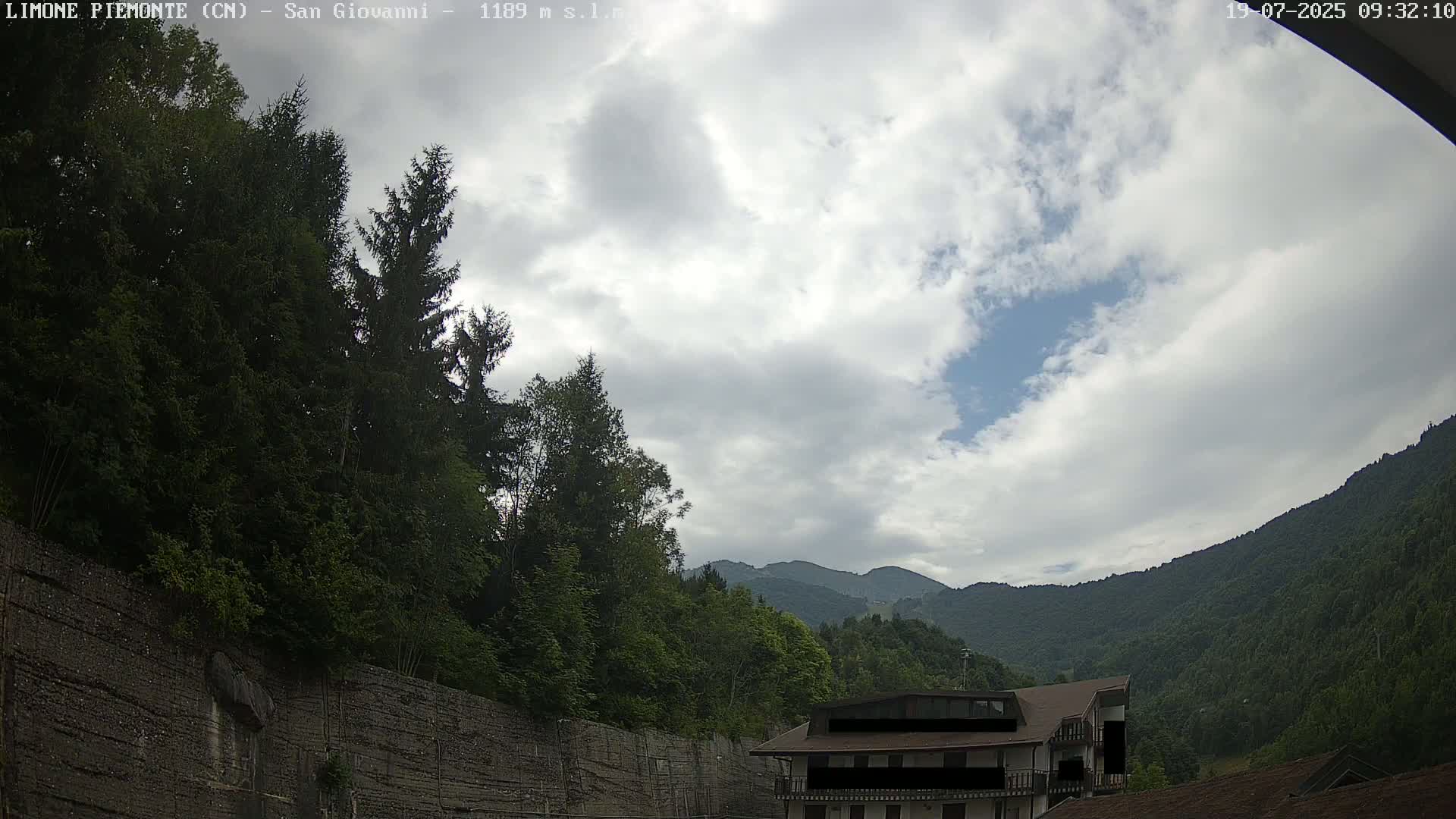 A partly cloudy sky overlooks a mountainous landscape with green trees and a building near a retaining wall.