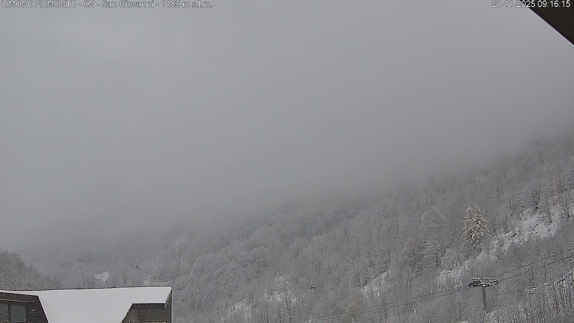 A snow-covered mountain landscape, dense with frosted trees and featuring a partially visible building and ski lift pole, is shrouded in heavy overcast skies or fog.