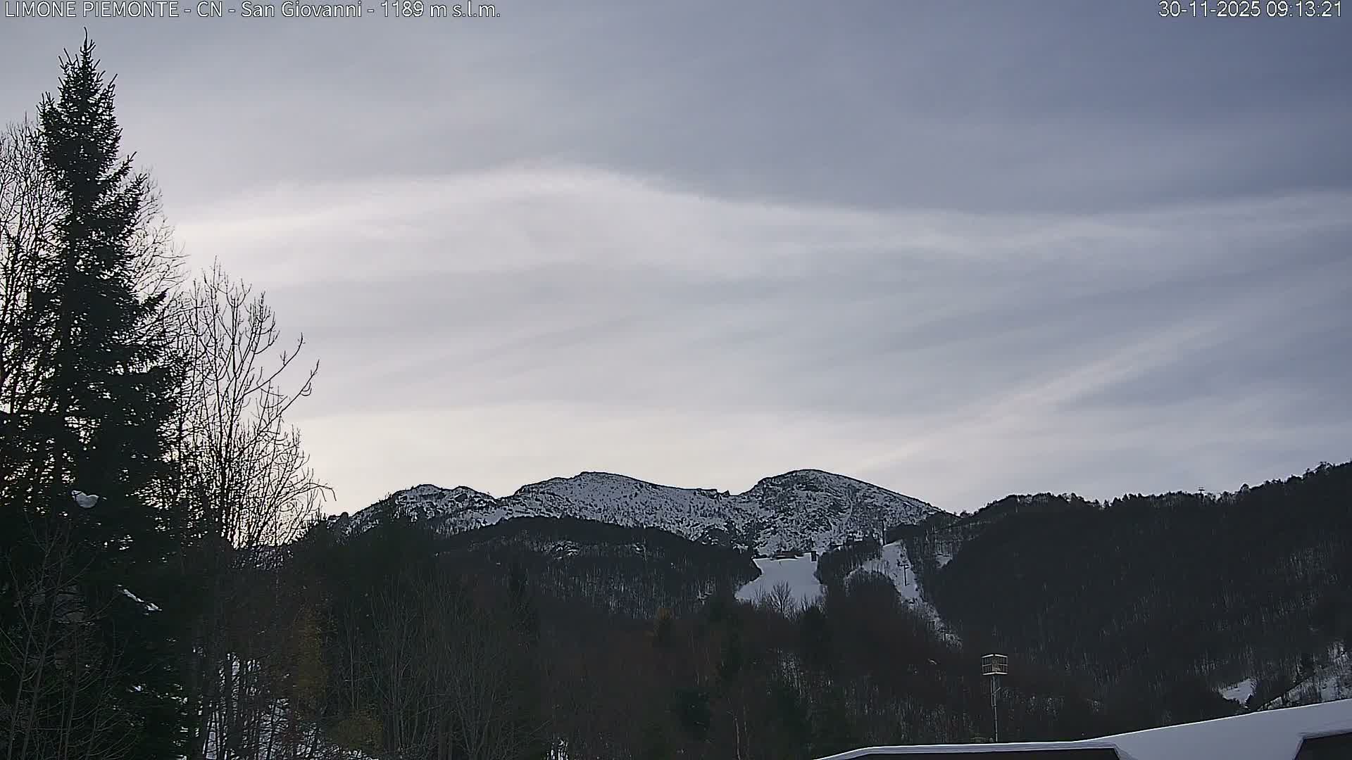 A winter mountain landscape is visible under an overcast sky, featuring snow-dusted peaks, a ski slope, and a mix of bare deciduous and evergreen trees on the lower slopes.