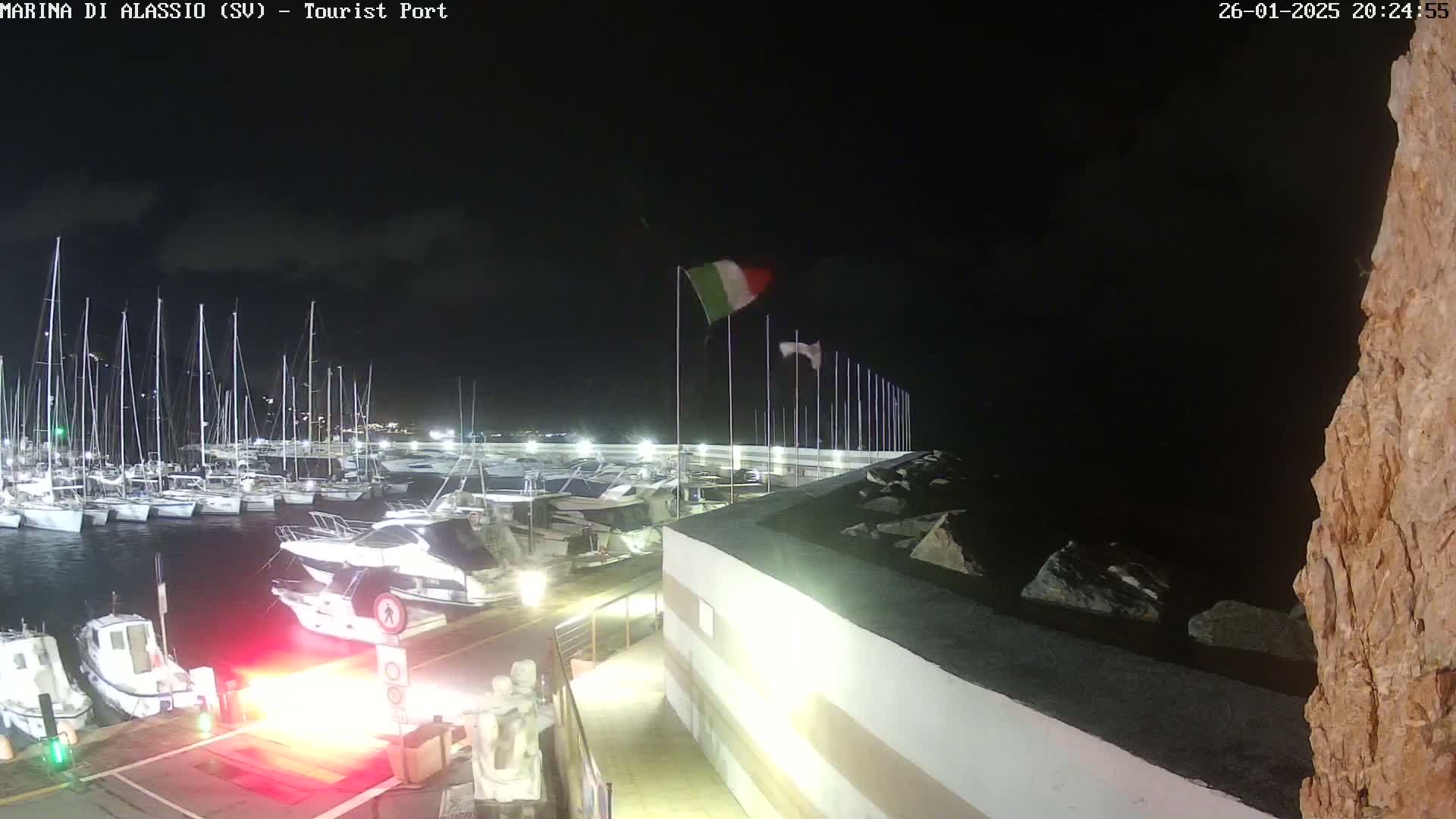 At night, a marina filled with sailboats and motorboats is illuminated under a partly cloudy sky.