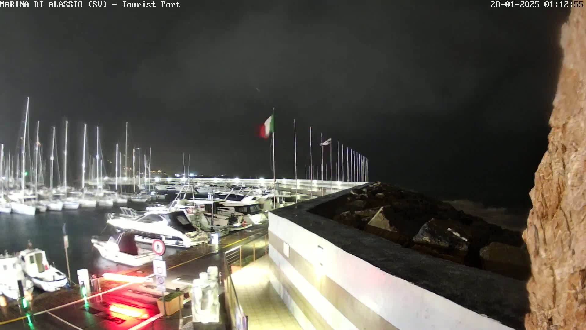 A nighttime view of a marina filled with sailboats and motorboats under a dark, cloudy sky, with rain on the wet dock.
