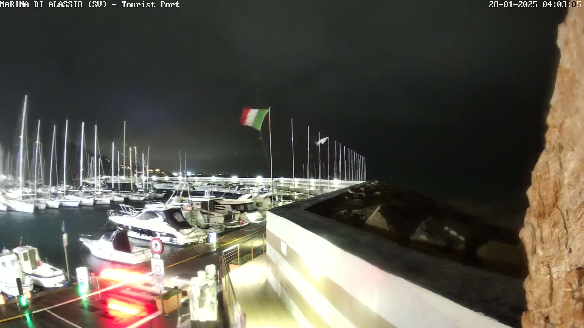 A nighttime view of a marina with numerous boats docked, under a dark, possibly stormy sky, with an Italian flag waving in the wind.