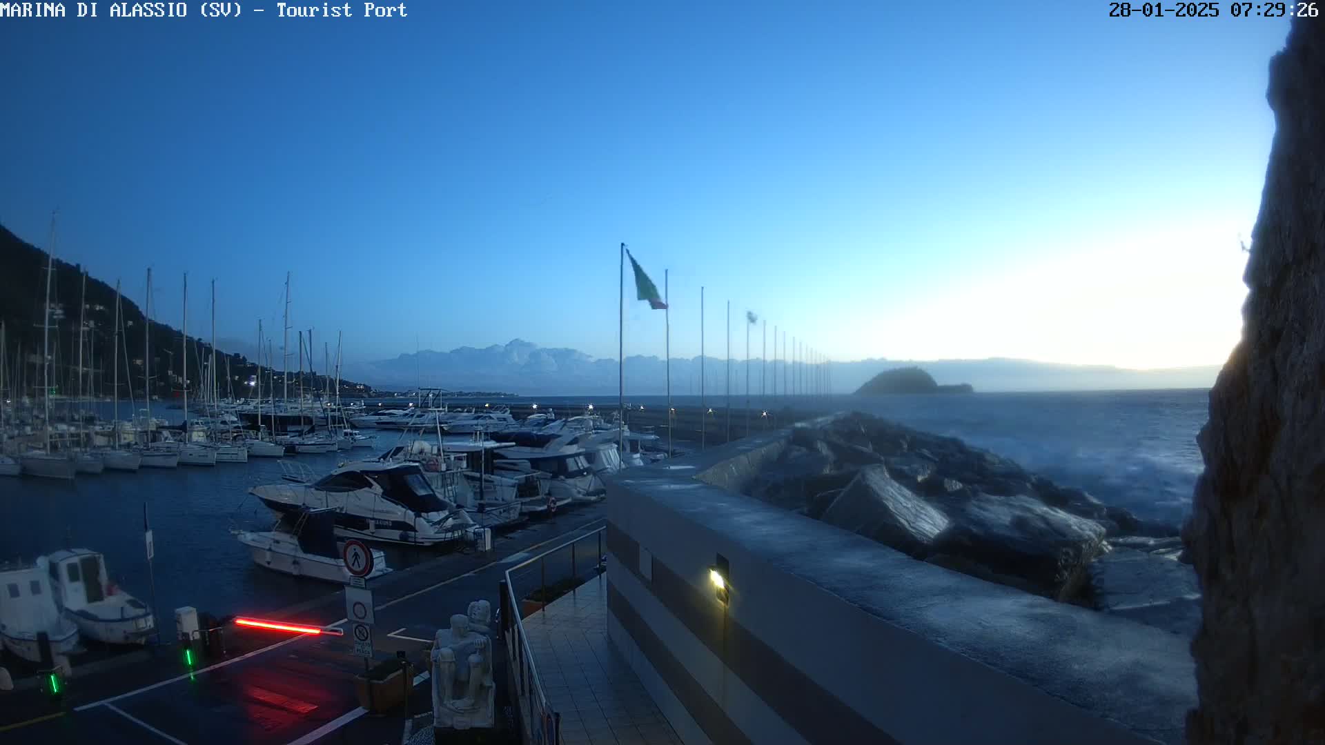 A marina filled with boats sits under a clear, twilight sky next to a rocky shoreline with choppy water.