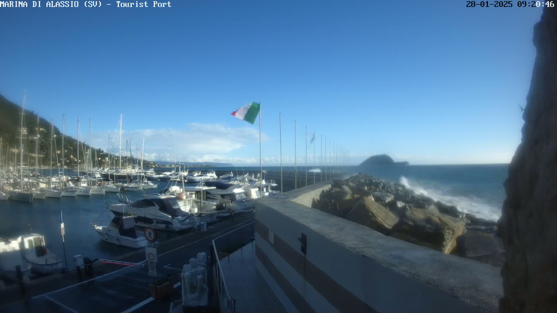 A harbor filled with boats is seen from a high vantage point on a mostly sunny day with some small clouds, and waves crashing against a rocky breakwater.