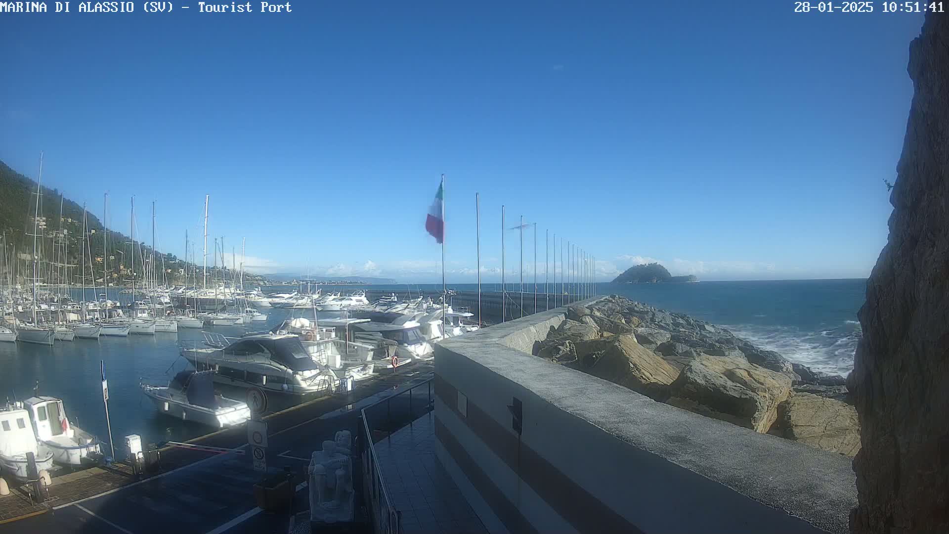 A marina filled with sailboats and motorboats is situated next to a rocky shoreline with a calm sea under a clear blue sky.