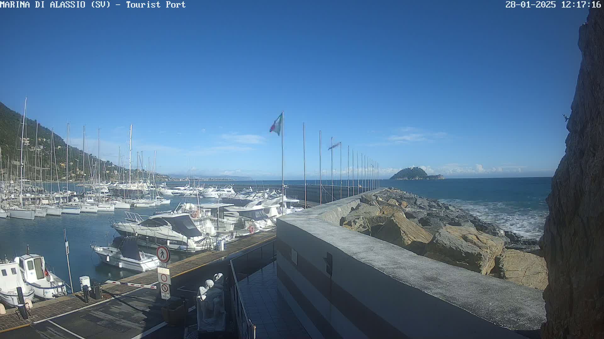 A marina filled with boats is situated on a calm, sunny day next to a rocky shoreline and a small island in the distance.