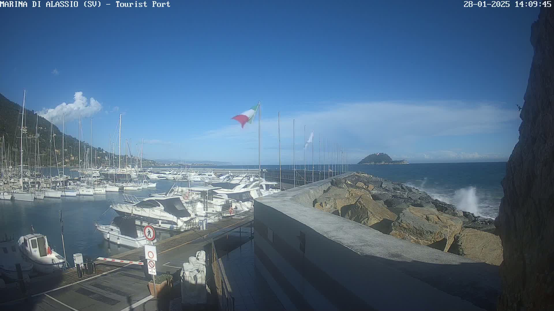 A marina filled with sailboats and motorboats is situated on a calm, sunny day next to a rocky shoreline with small waves breaking against it.