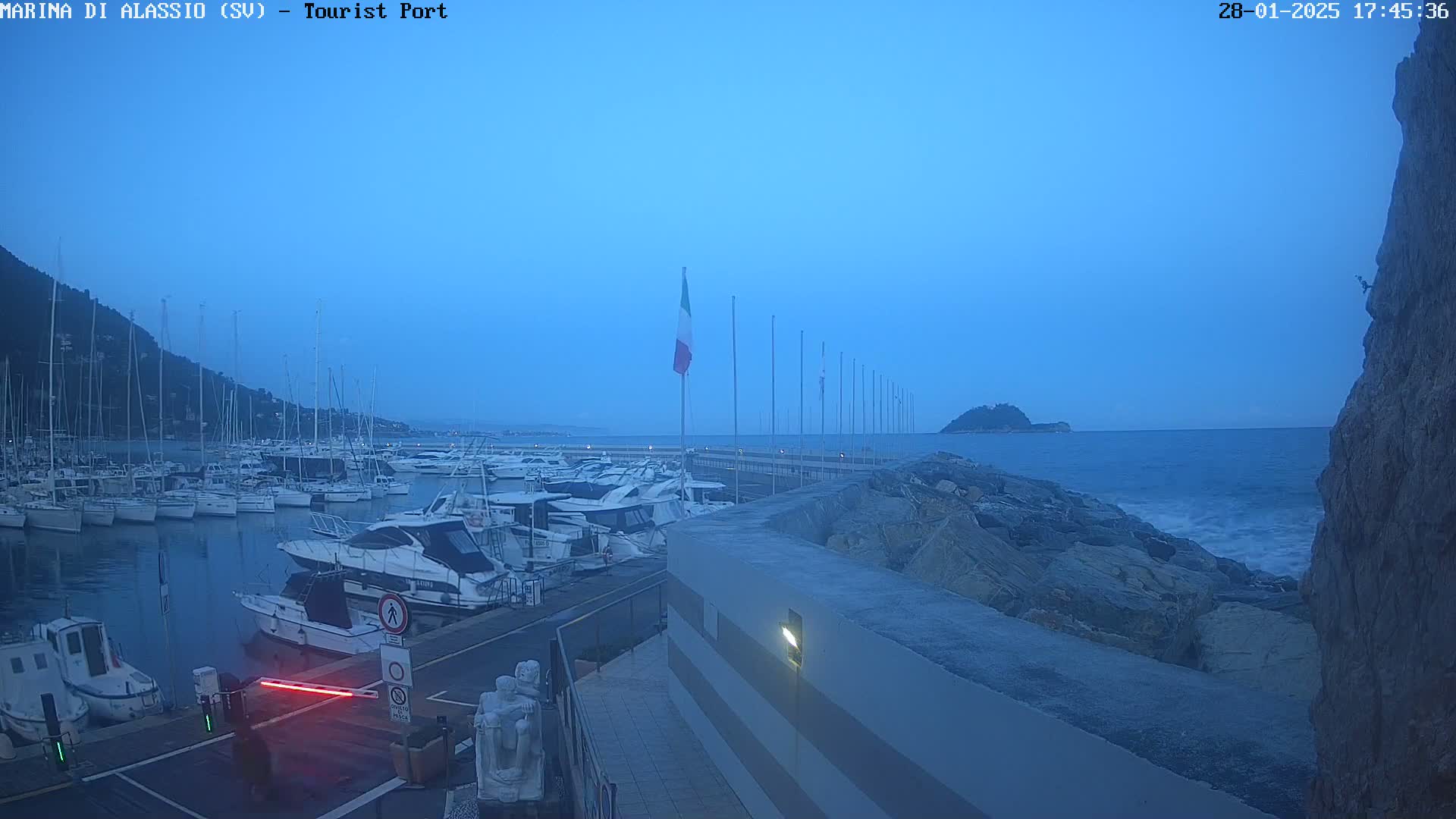 A marina filled with boats is seen at dusk under a clear, blue sky, with a rocky shoreline and calm waves visible in the distance.