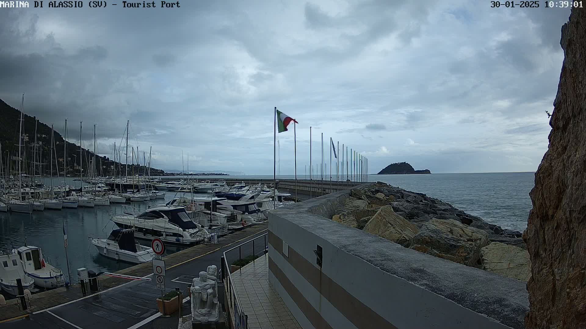 A marina filled with sailboats and motorboats is situated on a partly cloudy day next to a rocky shoreline and a small island in the distance.