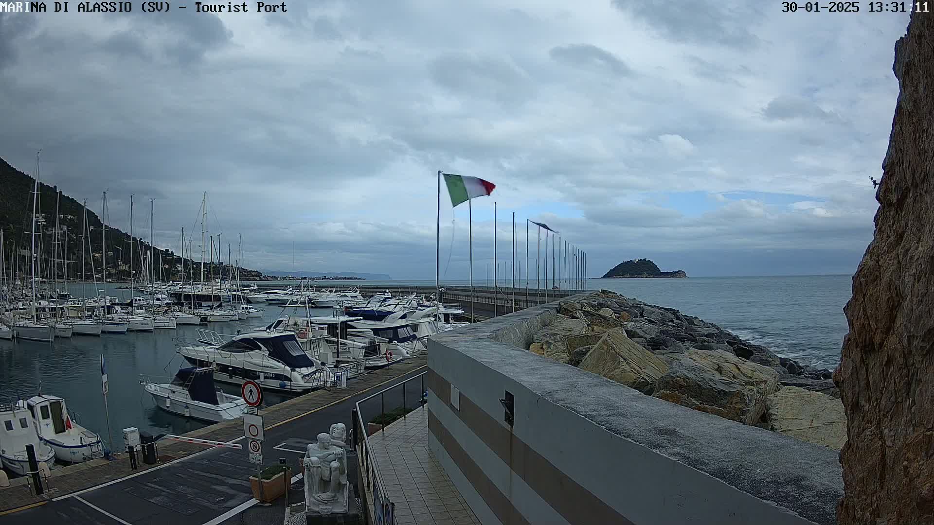 A harbor filled with sailboats and motorboats is seen under a cloudy sky, with a rocky breakwater separating the harbor from the open sea.