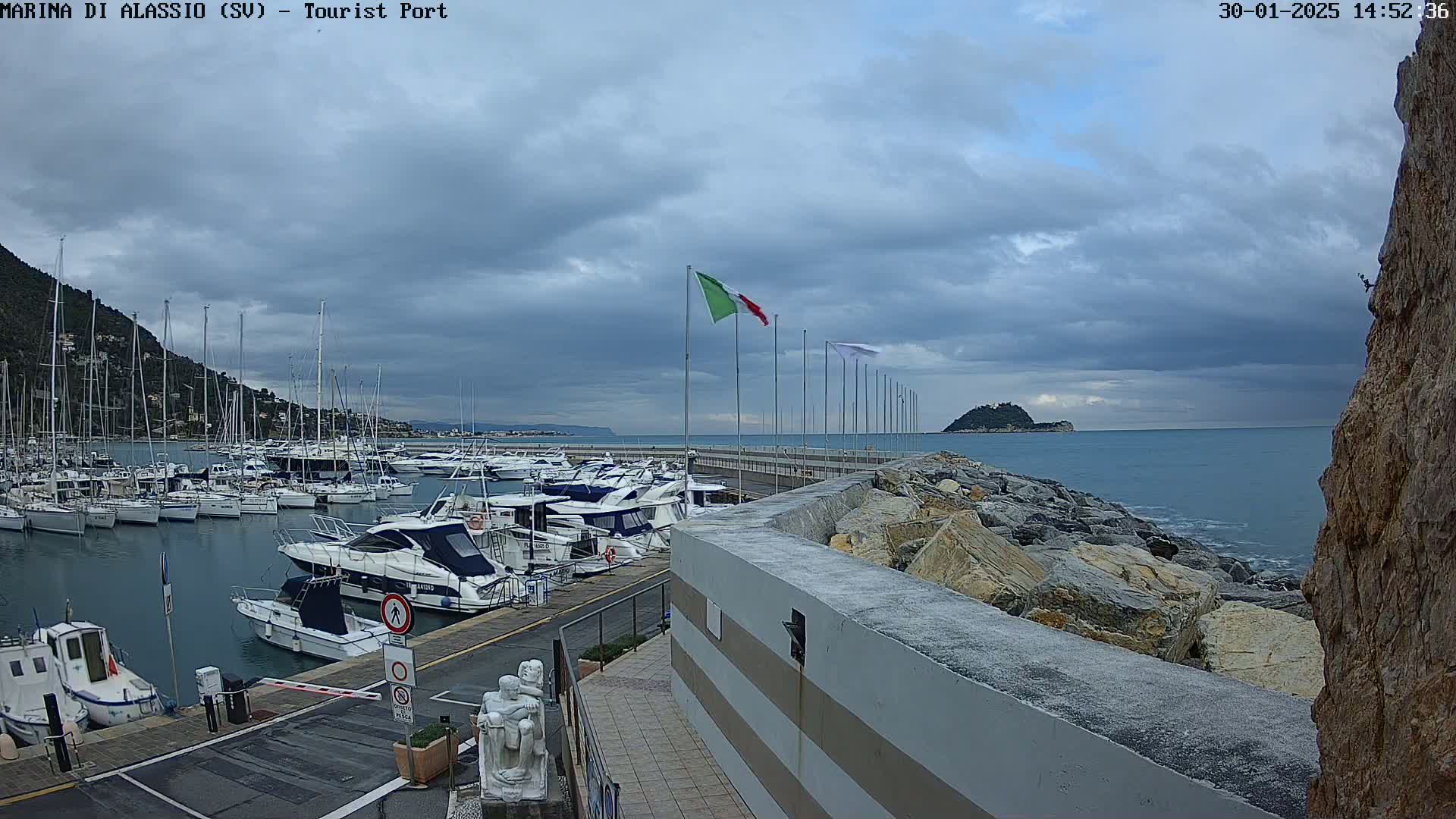 A marina filled with boats is situated next to a rocky shoreline under a cloudy sky.
