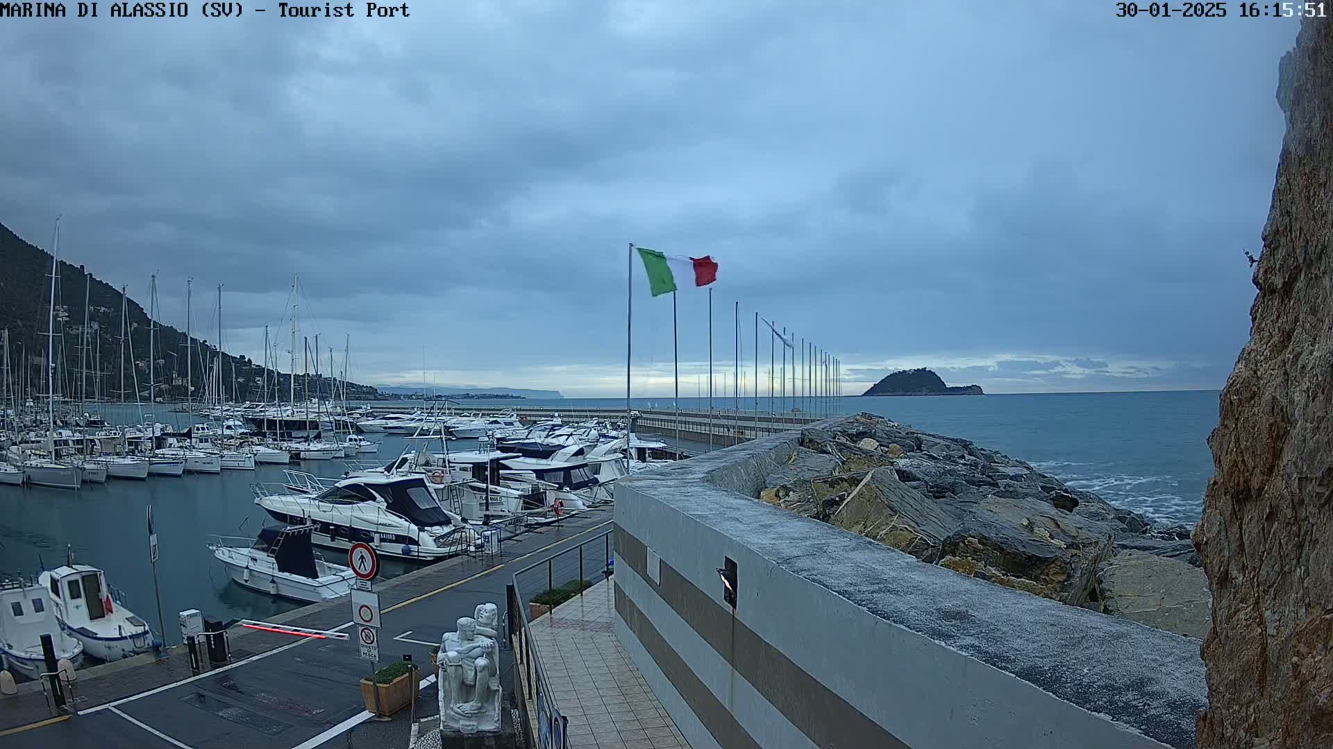 A harbor filled with numerous boats is situated under a cloudy sky, with a rocky shoreline and calm ocean visible in the background.