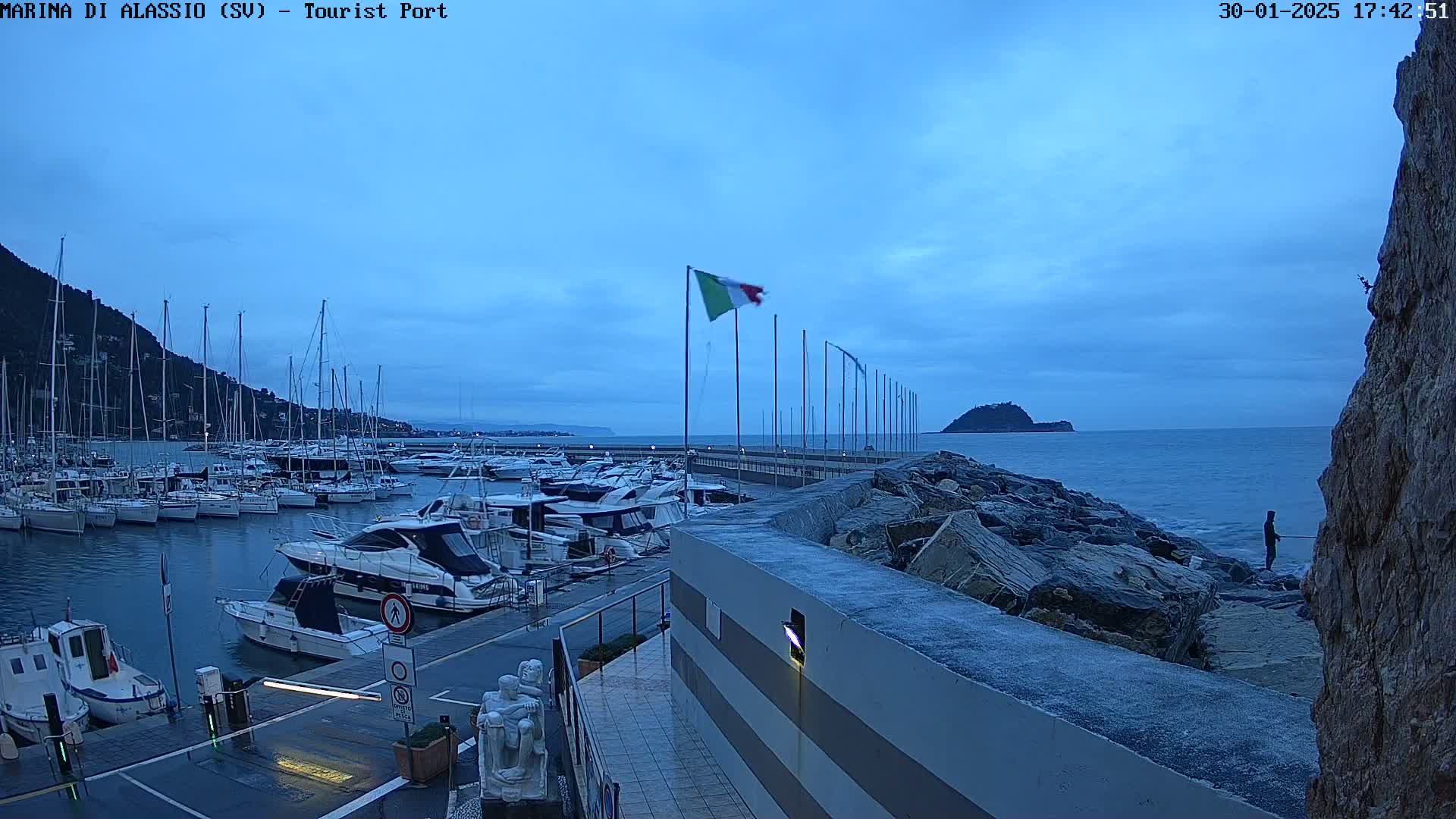 A marina filled with sailboats and motorboats is situated next to a rocky shoreline under a twilight sky.