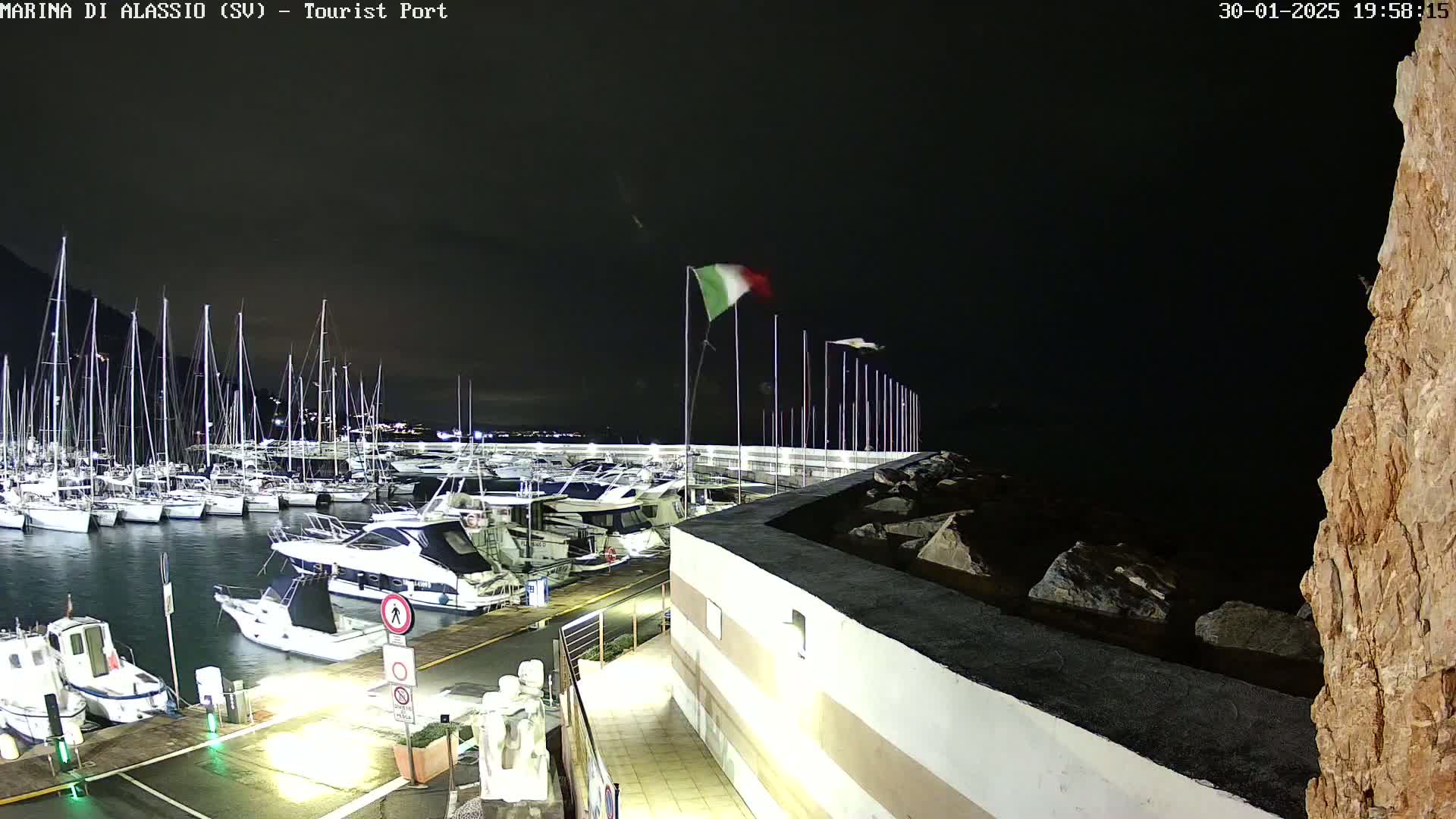 A nighttime view of a harbor filled with sailboats and motorboats, with an Italian flag waving in the wind.