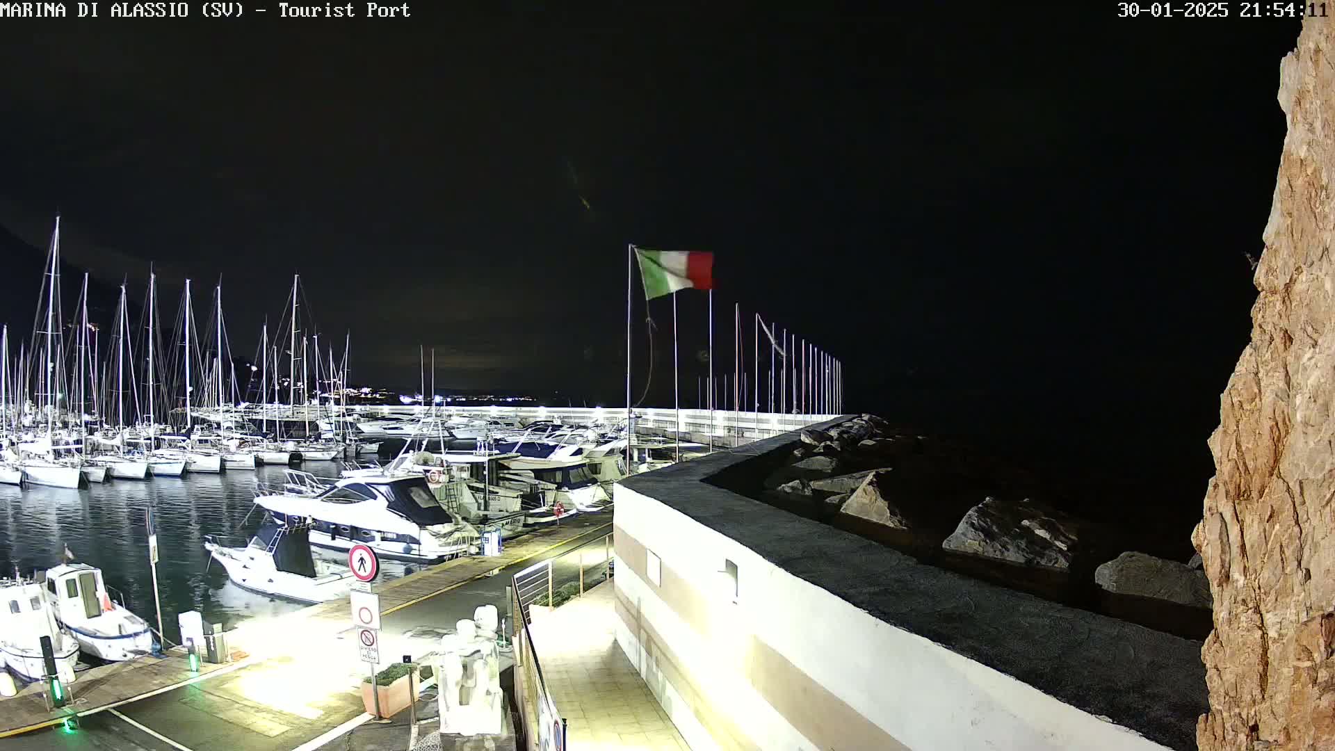A nighttime view of a marina filled with sailboats and motorboats, under a dark sky with an Italian flag waving.