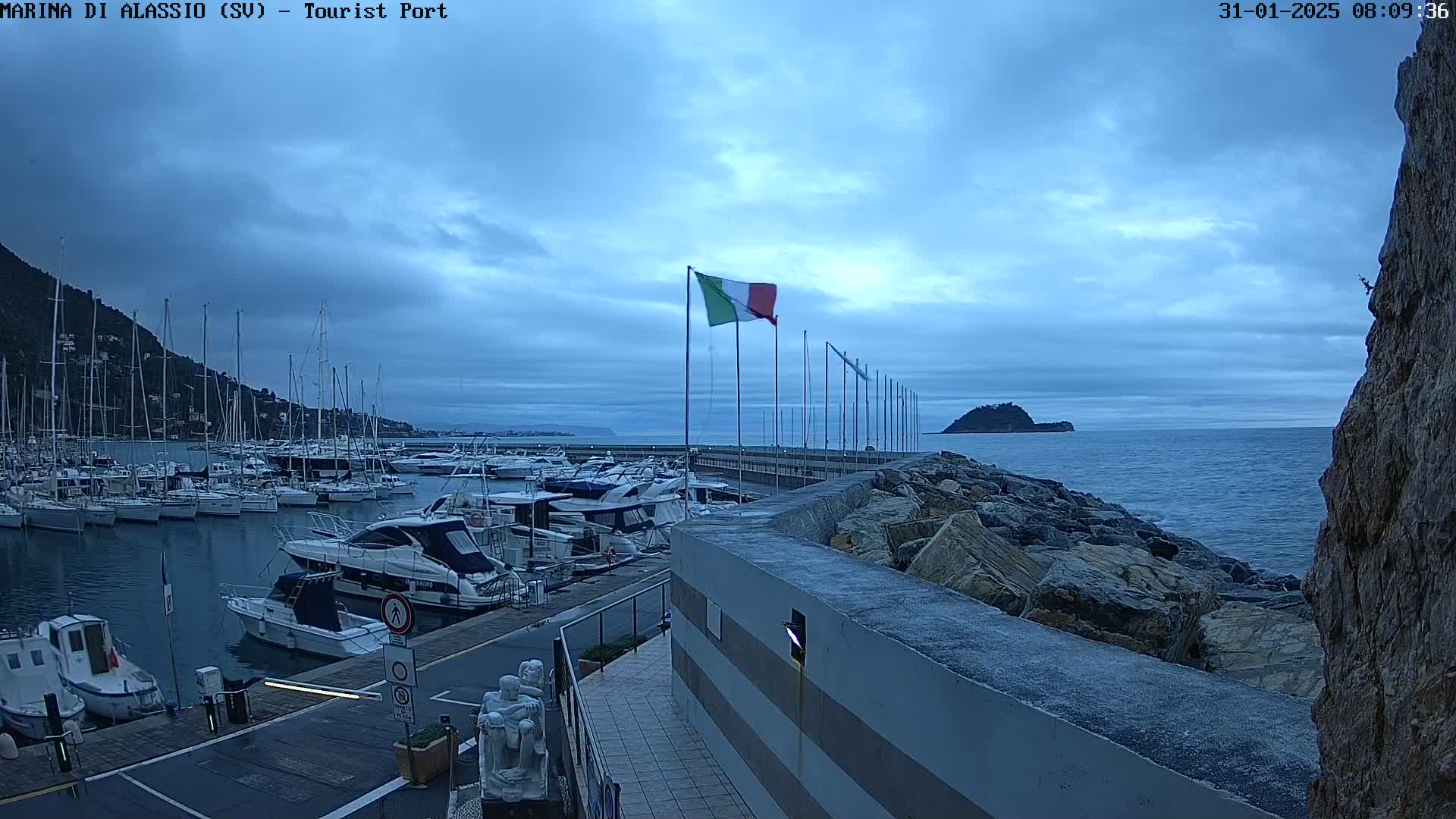 A marina filled with sailboats and motorboats is situated on a calm, overcast day next to a rocky coastline with an island in the distance.