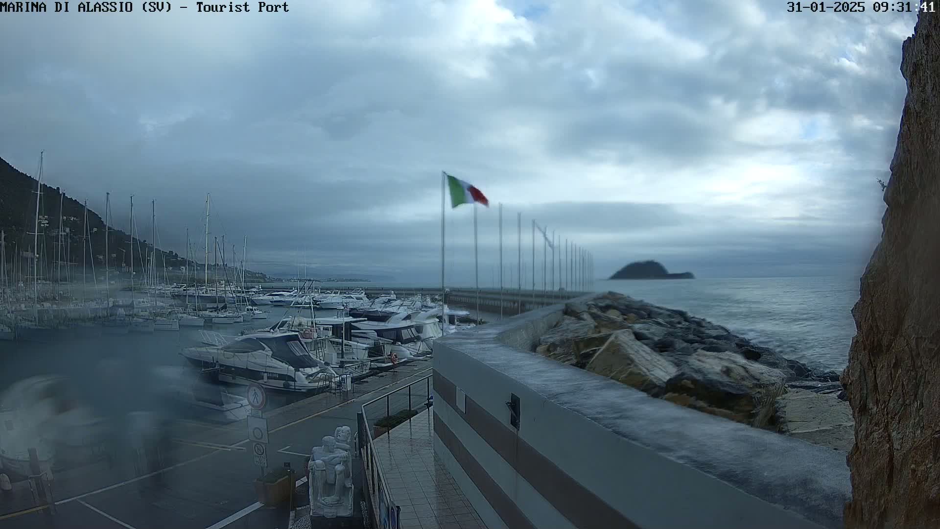 A marina filled with boats is shown under a cloudy sky, with a calm sea visible in the background.