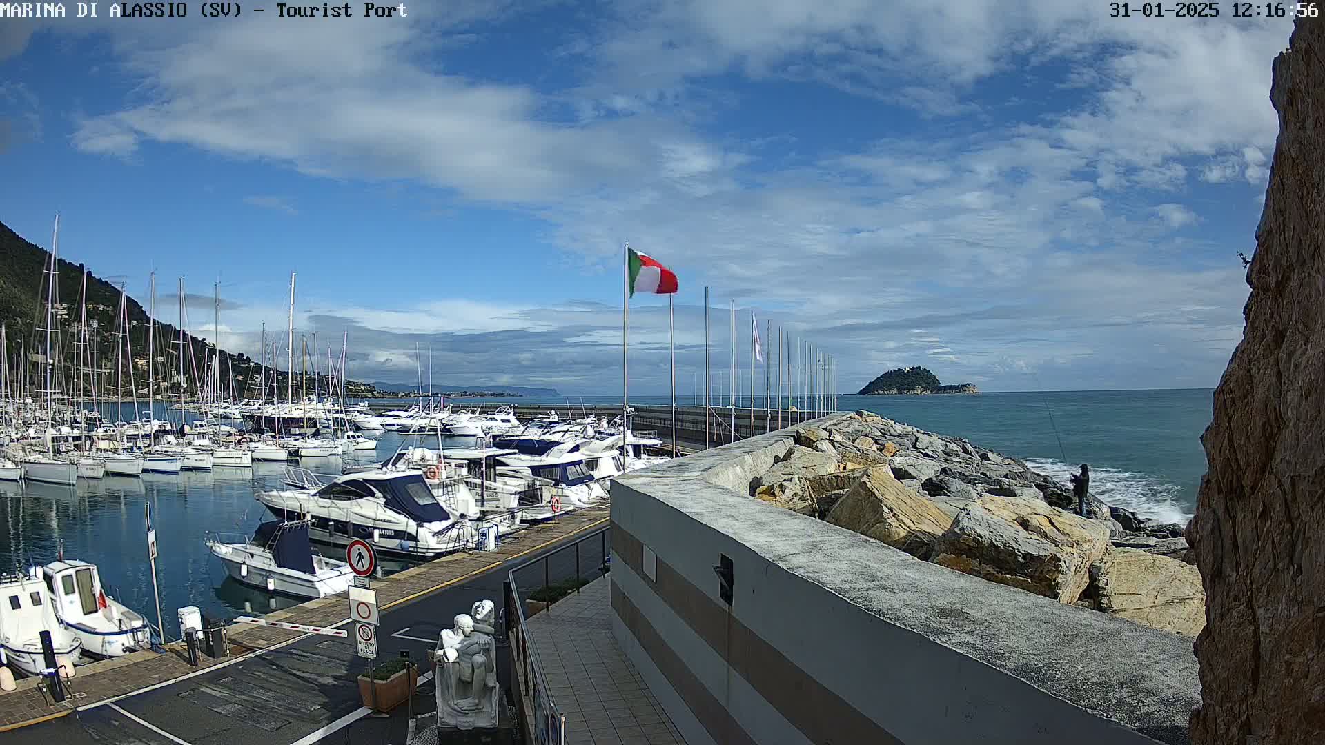 A harbor filled with numerous boats under a mostly sunny sky, with a person fishing from a rocky outcrop in the foreground.
