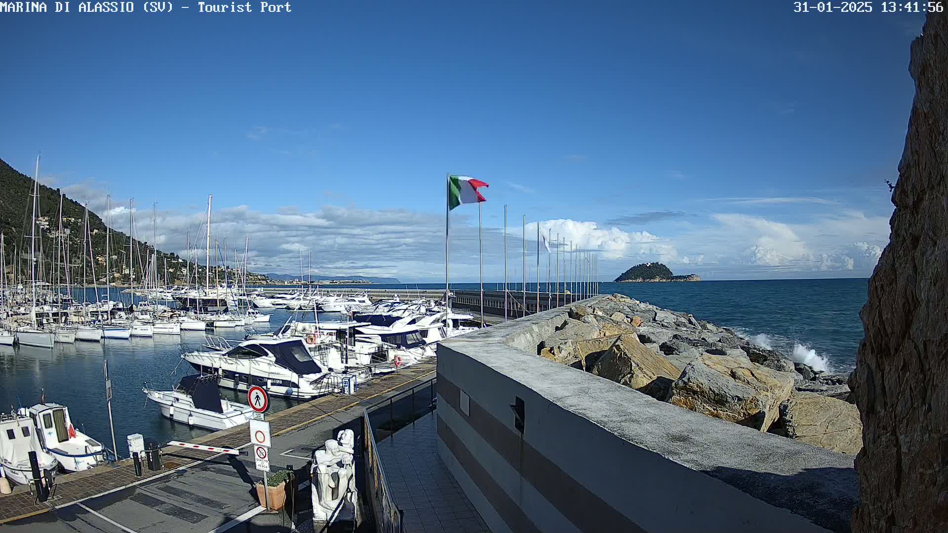 A harbor filled with boats is shown on a partly cloudy day, with a rocky breakwater separating the harbor from the open sea.