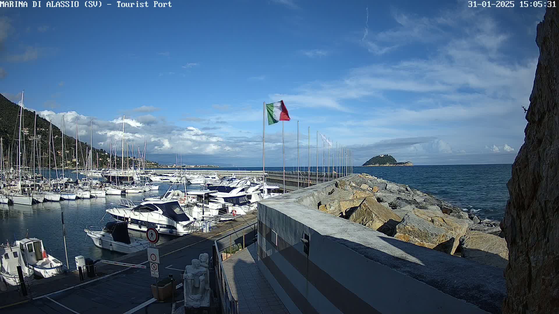 A marina filled with sailboats and motorboats is situated beside a rocky shore under a partly cloudy sky.
