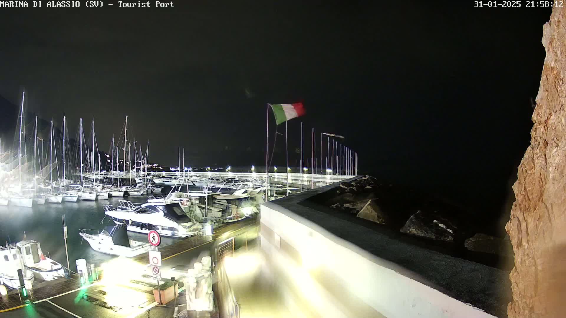 A nighttime view of a marina filled with boats, an Italian flag waving in the wind, and a rocky cliffside to the right.