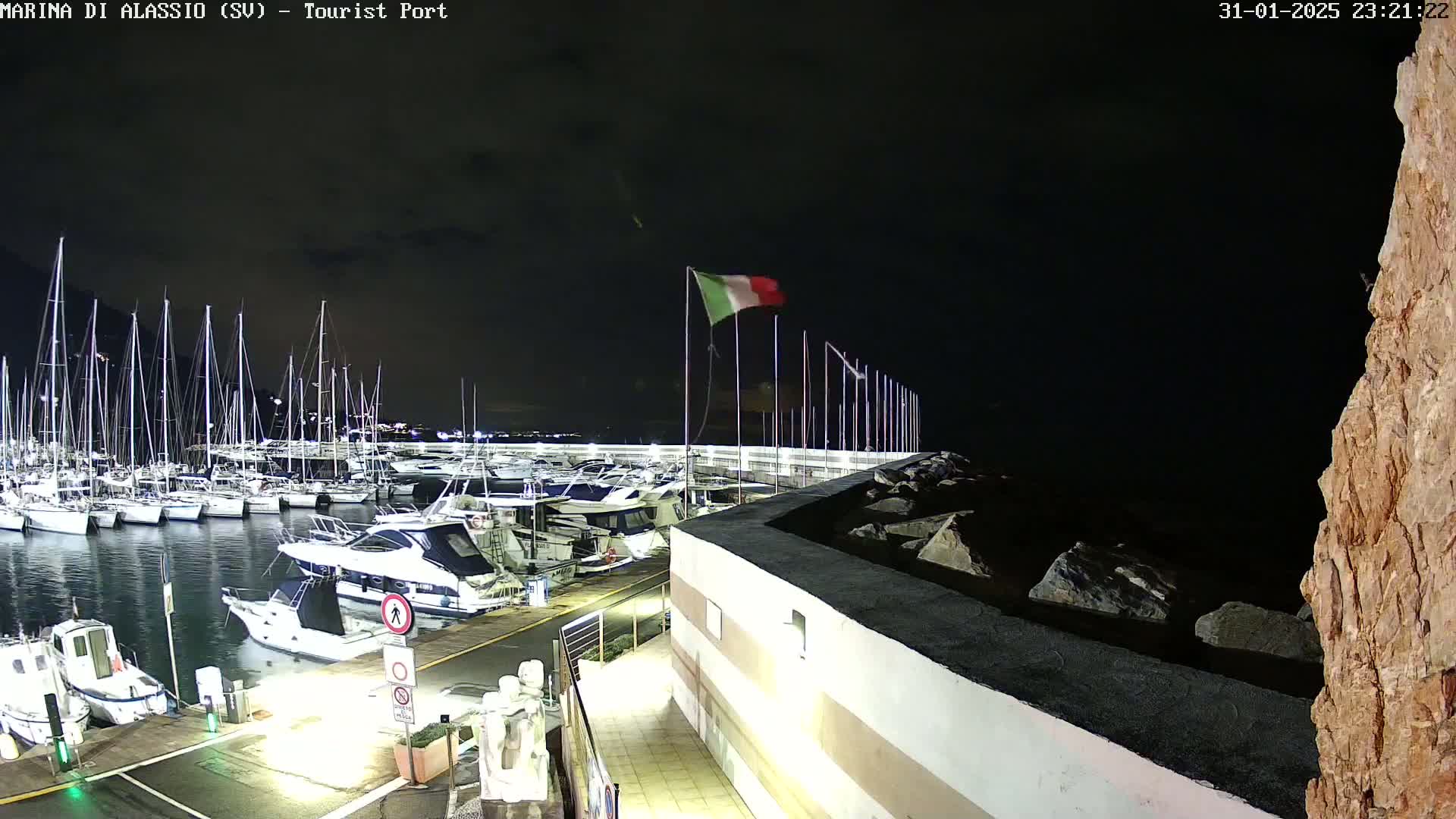 A nighttime view of a marina filled with boats, a rocky shoreline, and an Italian flag waving gently under a dark sky.
