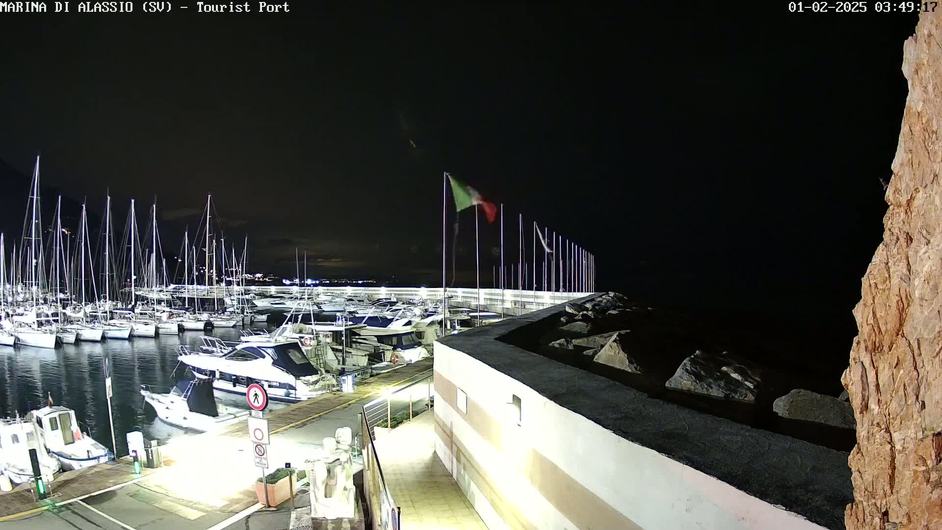 A nighttime view of a marina filled with boats, a rocky shore, and an Italian flag waving in the dark.