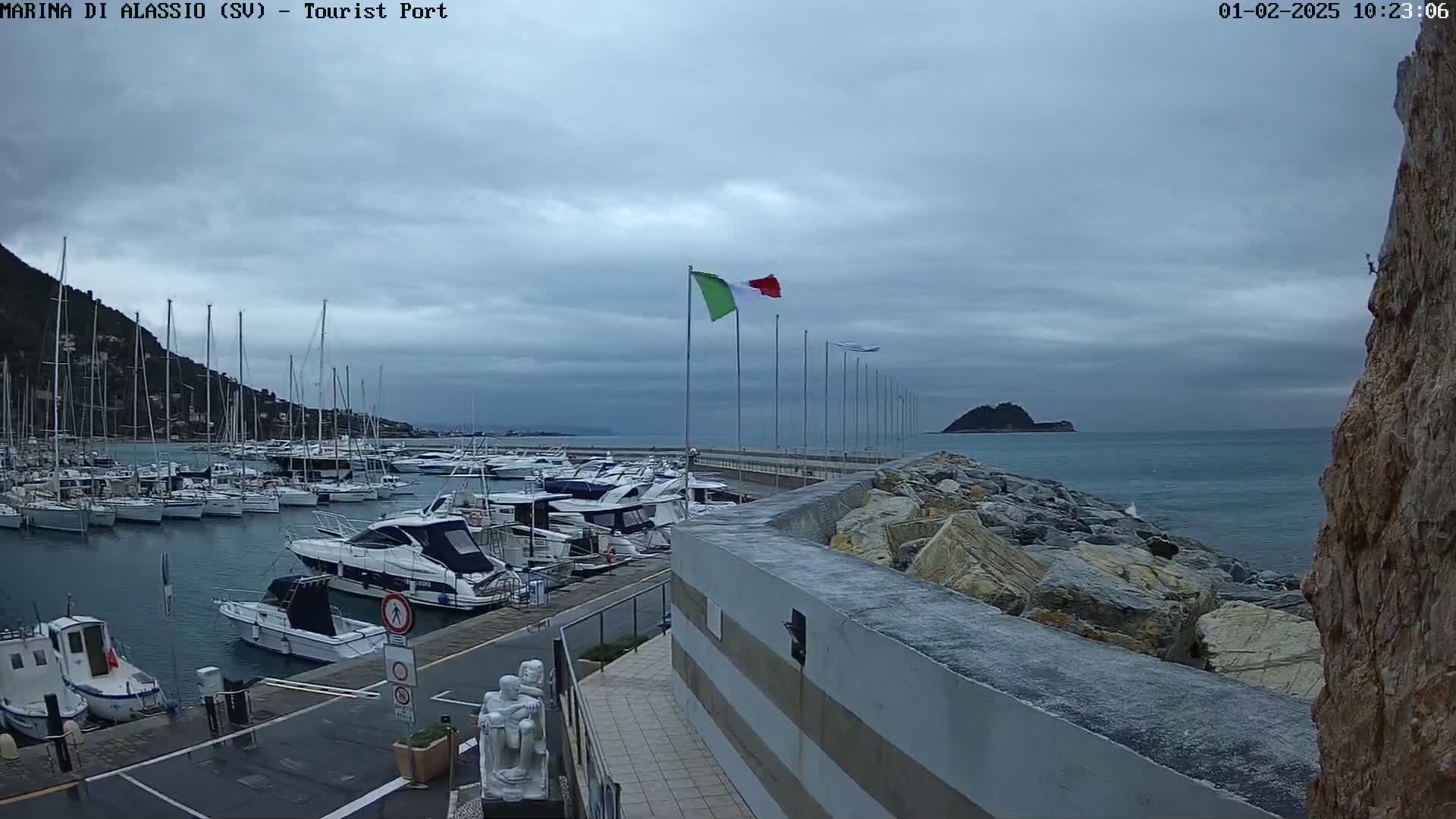 A harbor filled with numerous boats is situated under a cloudy sky, with a rocky breakwater extending into a calm sea.